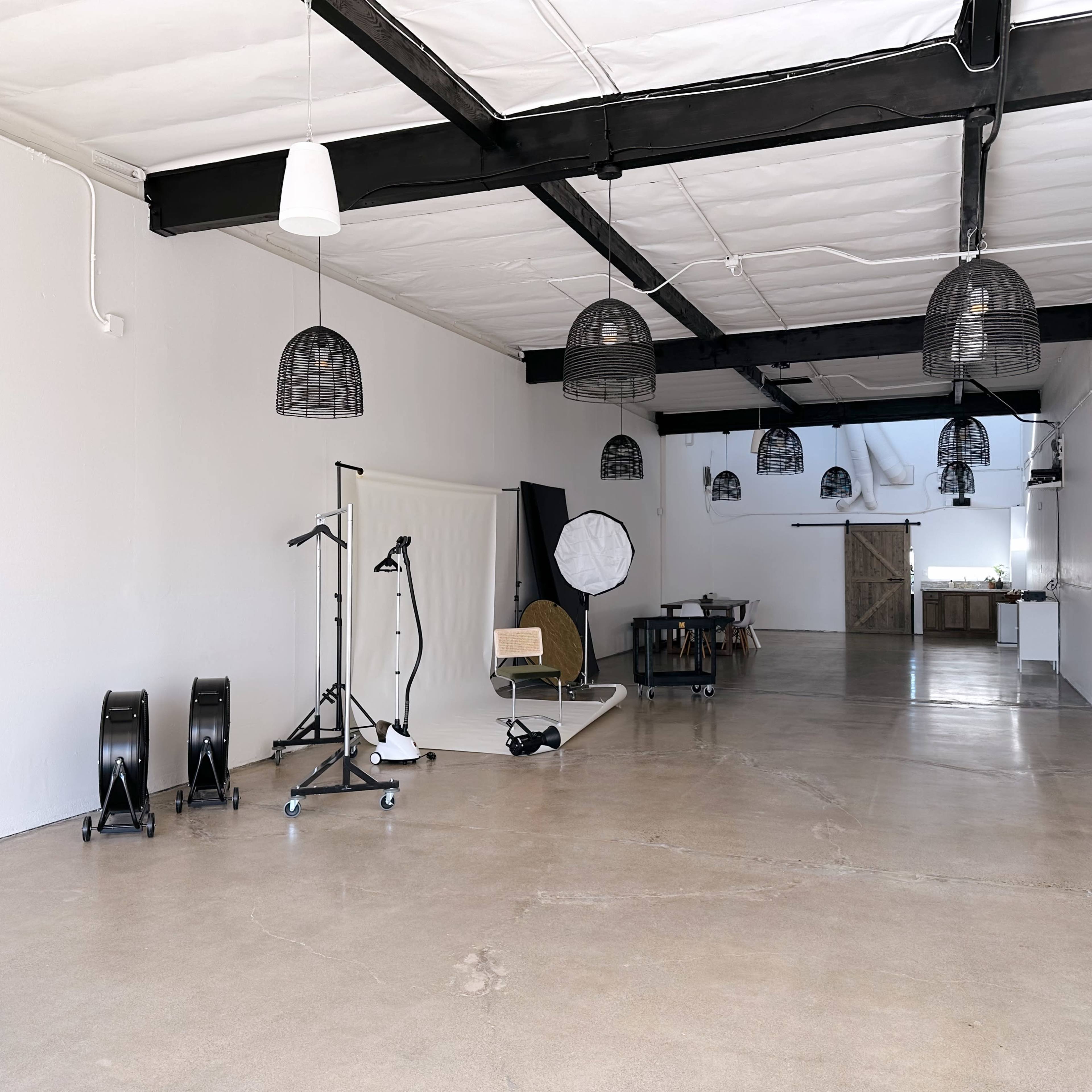 A spacious, minimalistic studio with concrete flooring and several hanging light fixtures, empty except for photography equipment and props arranged along the left wall.