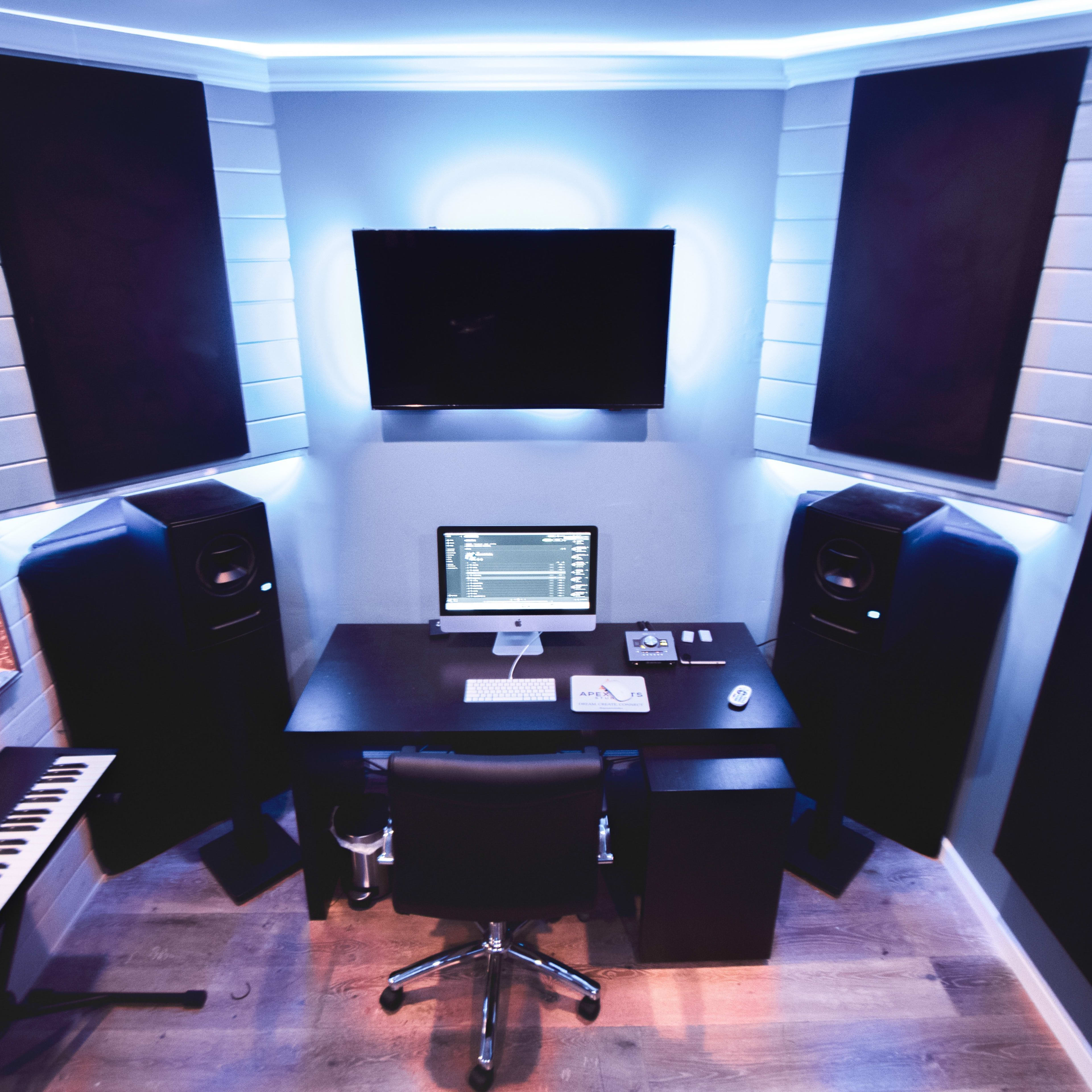 A modern home music studio with a computer desk, two large speakers, a keyboard, and a wall-mounted TV in a corner setup.