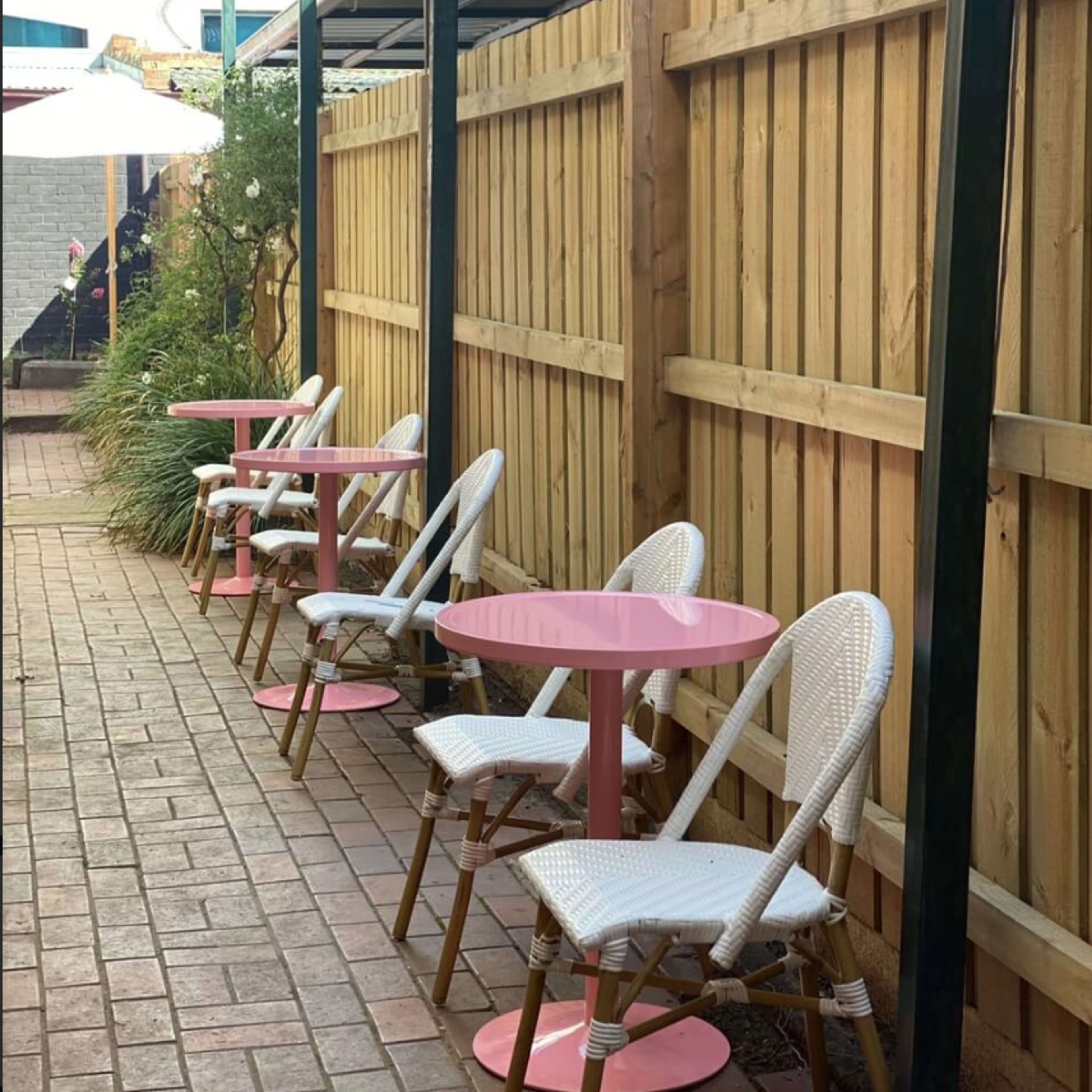 A row of white chairs with pink tables is placed against a wooden fence in an outdoor setting.
