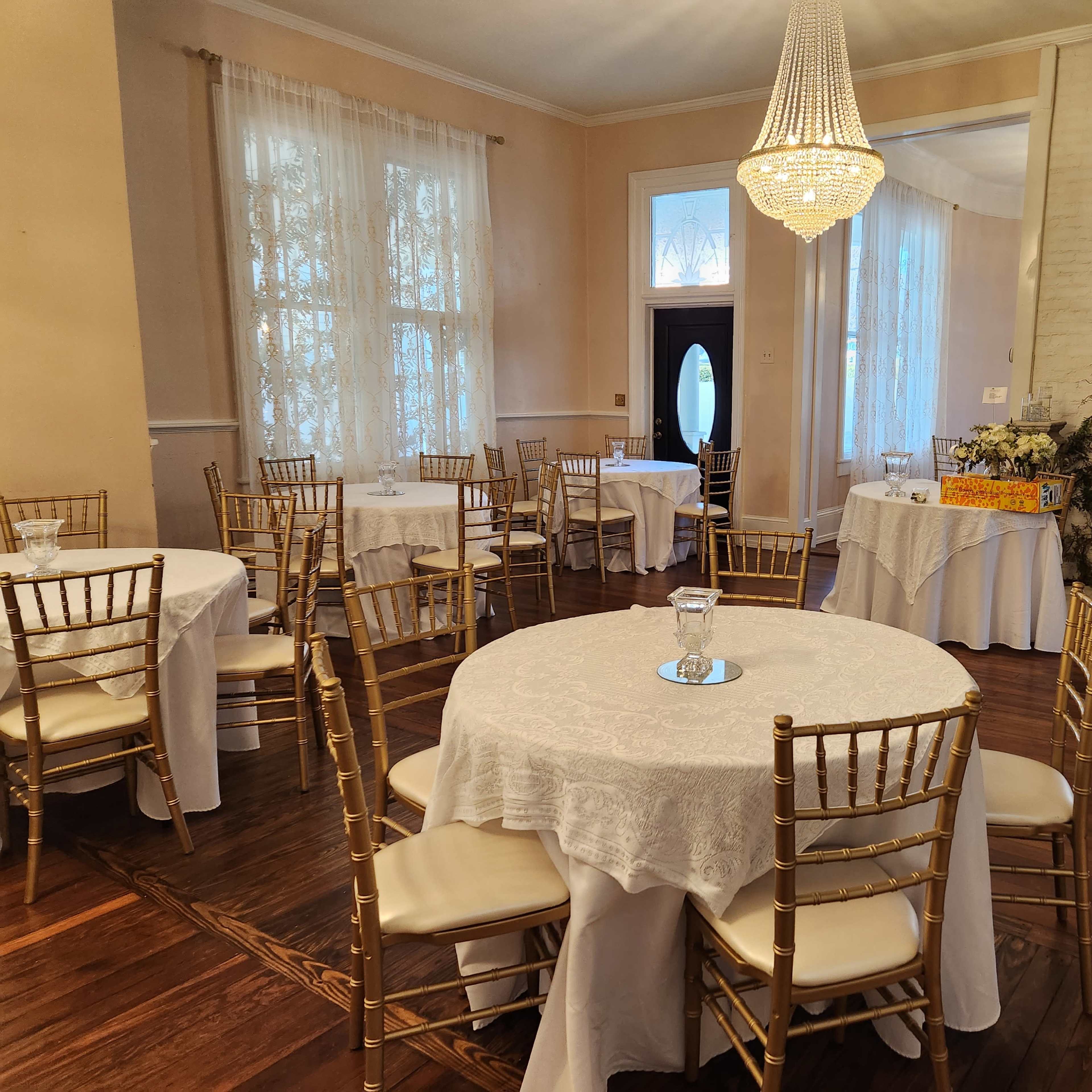 The scene shows a decorated room with round tables covered in white tablecloths and gold chairs, featuring a chandelier and large windows with sheer curtains.