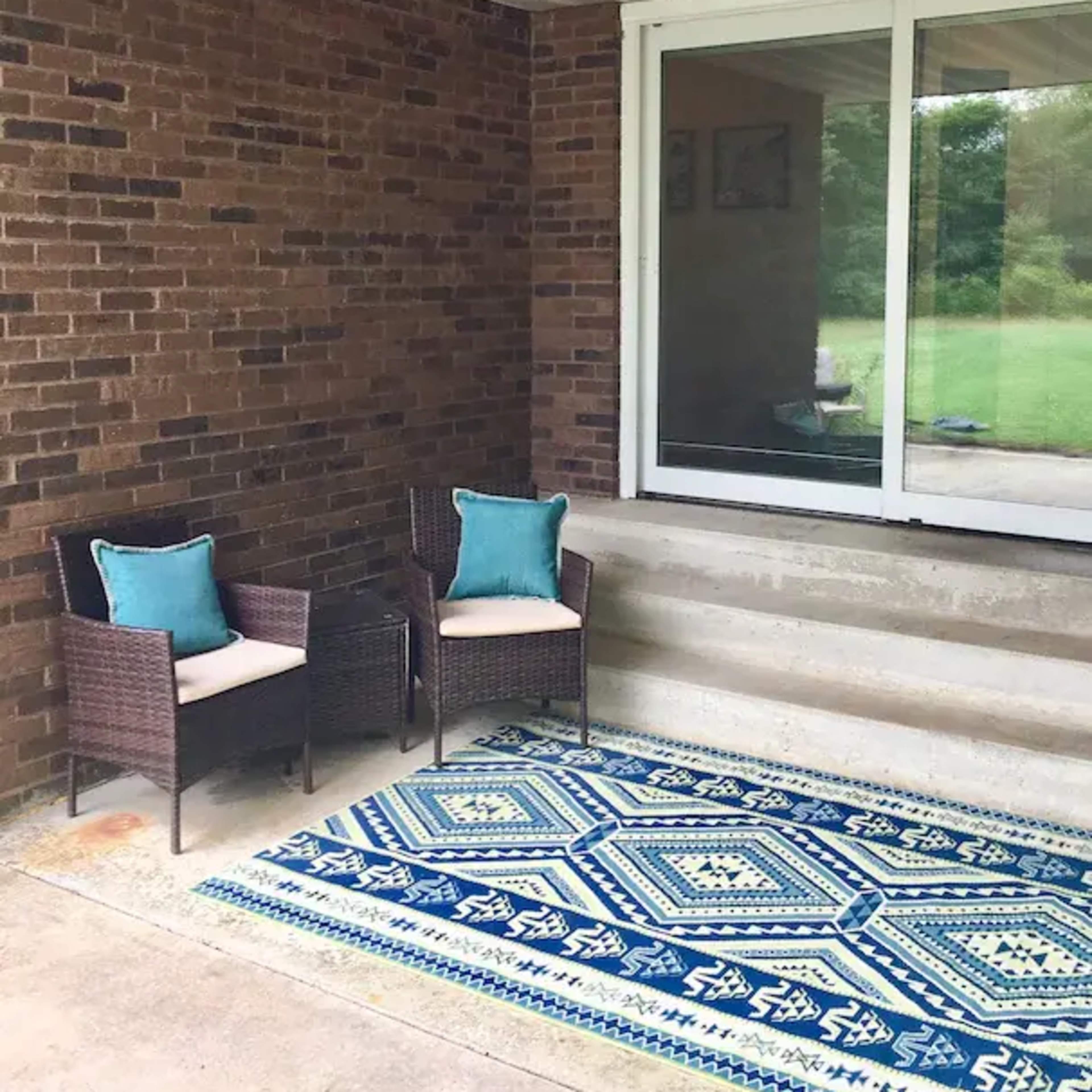 Two wicker chairs with teal cushions are positioned on a patterned blue area rug in front of a sliding glass door, with a brick wall and steps leading up to the entrance.