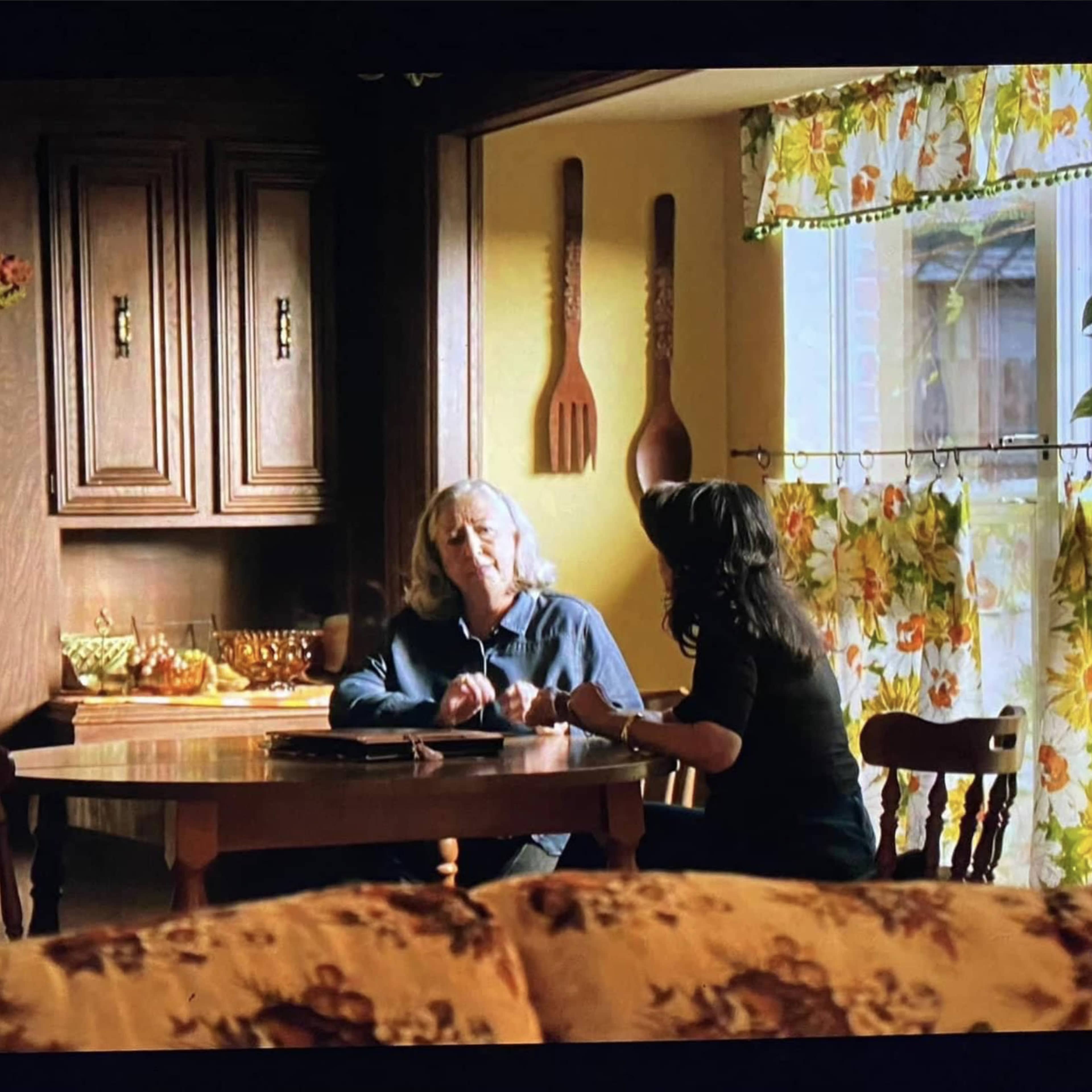 Two women are seated at a table in a warmly lit kitchen, engaged in conversation.