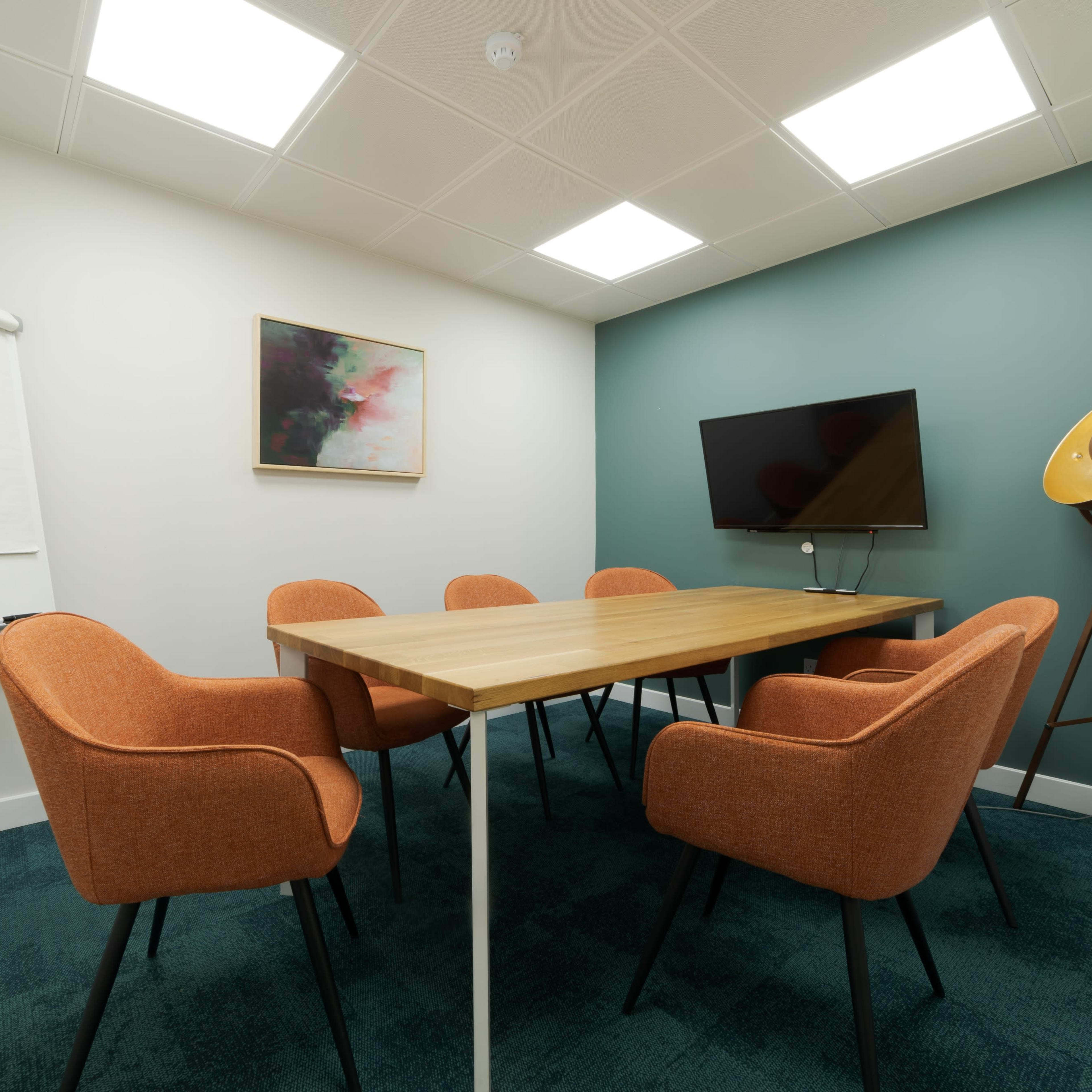 A conference room with a wooden table surrounded by six orange chairs, a whiteboard, a television mounted on the wall, and a decorative floor lamp.