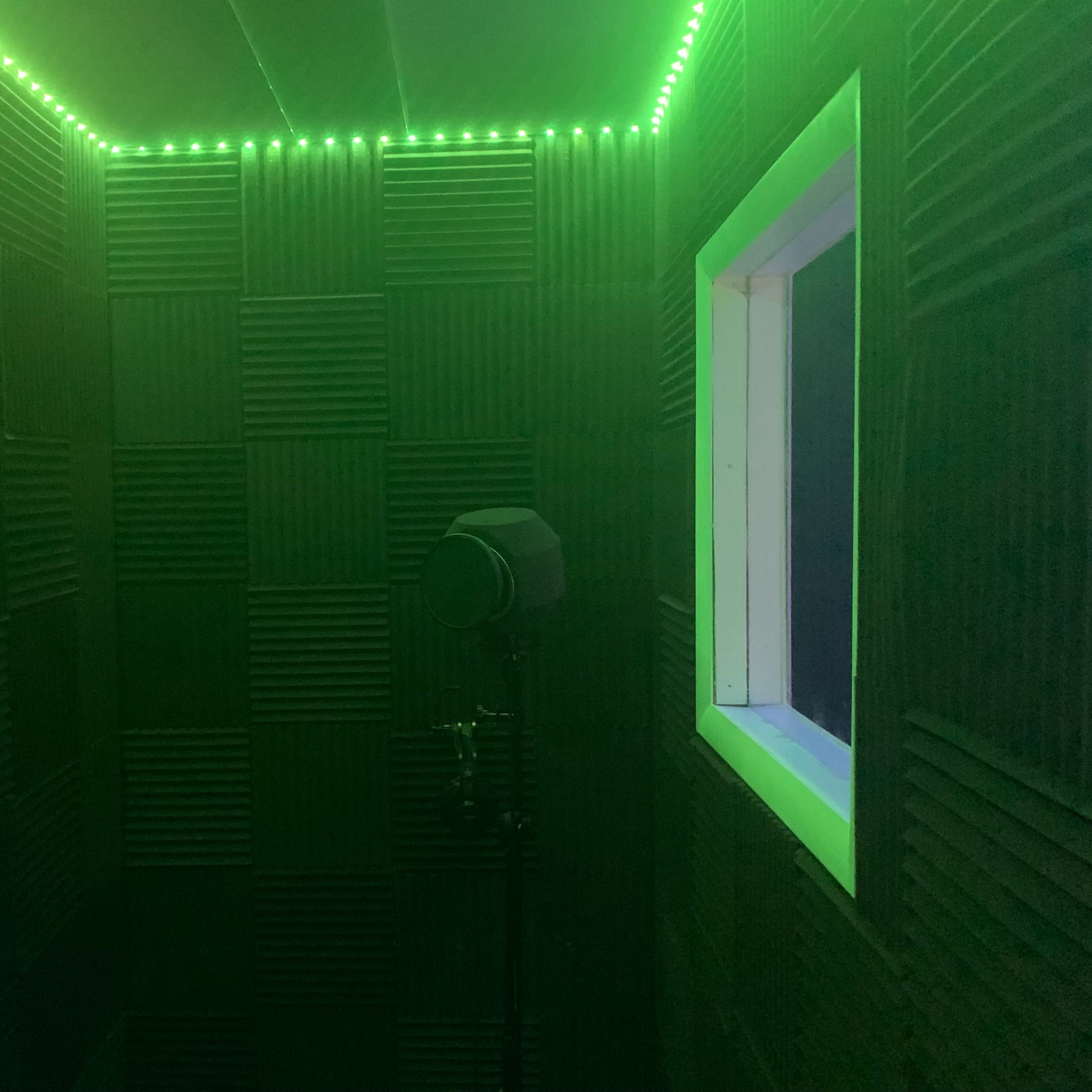 A small room with soundproof walls, illuminated by green LED lights, featuring a microphone stand and a window.