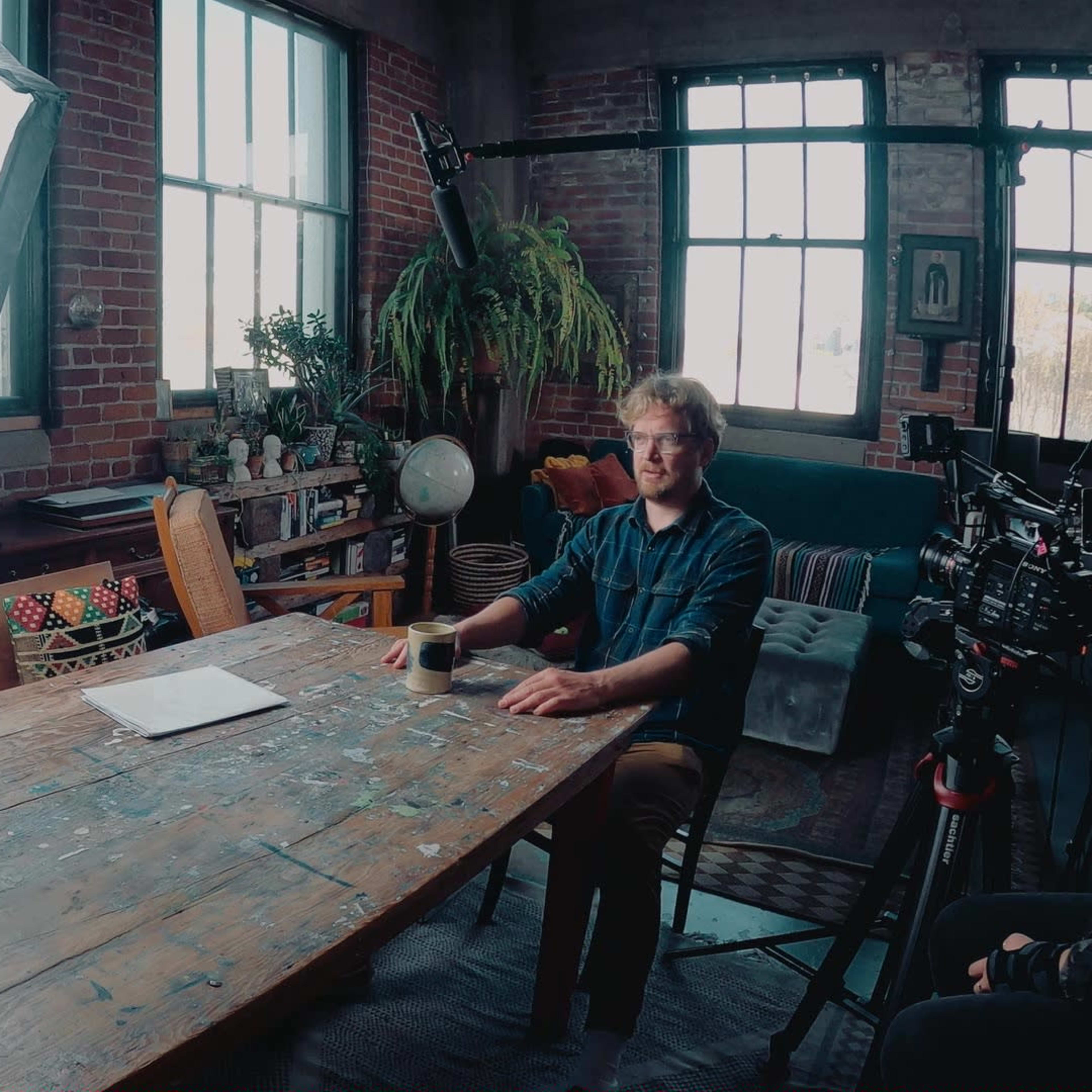 A man sits at a large, weathered wooden table in a studio filled with plants and equipment, with large windows letting in natural light.