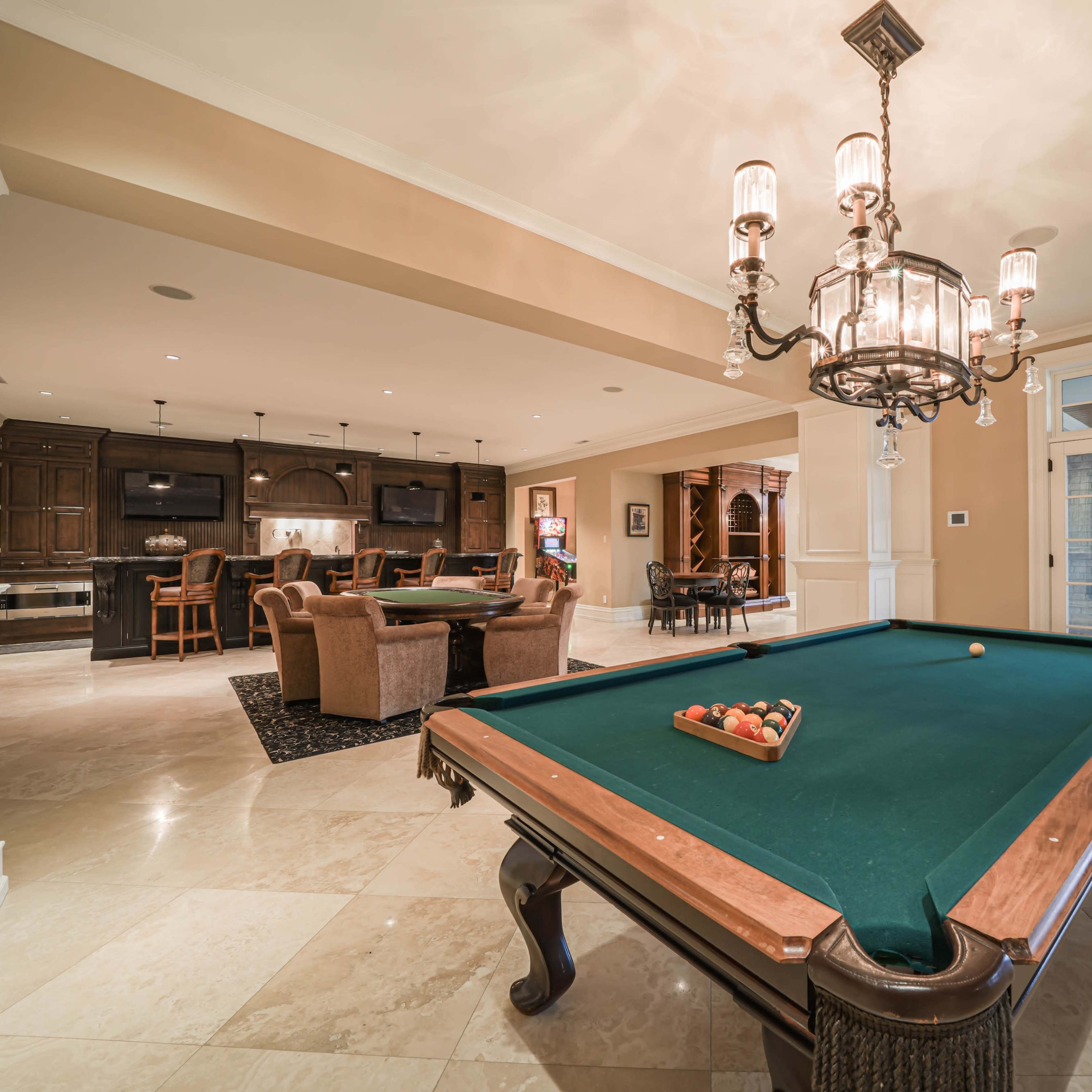A spacious game room featuring a green billiards table in the foreground, with a dining area and a kitchen in the background.