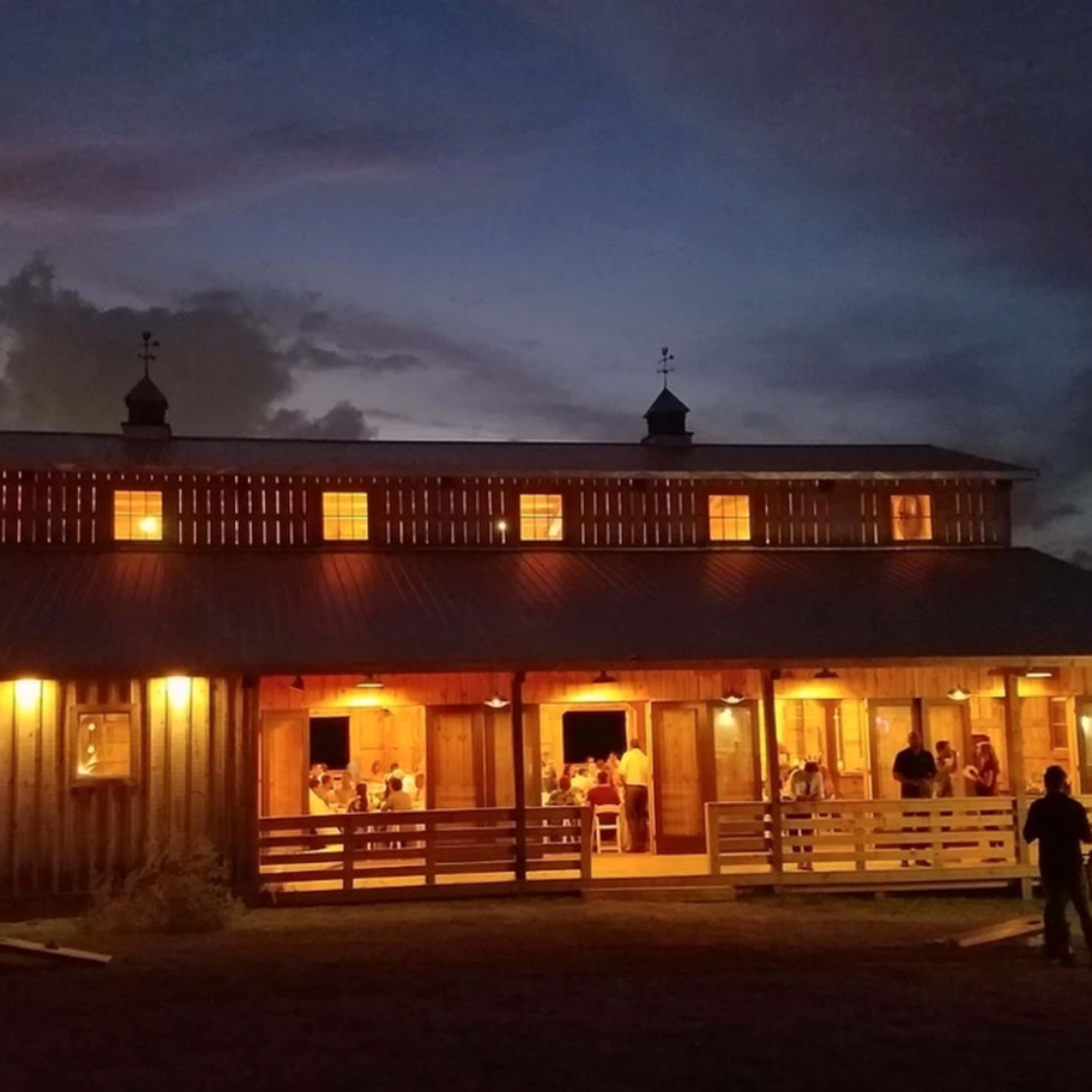 A wooden barn illuminated by warm lights stands against a twilight sky with people gathered on its porch.