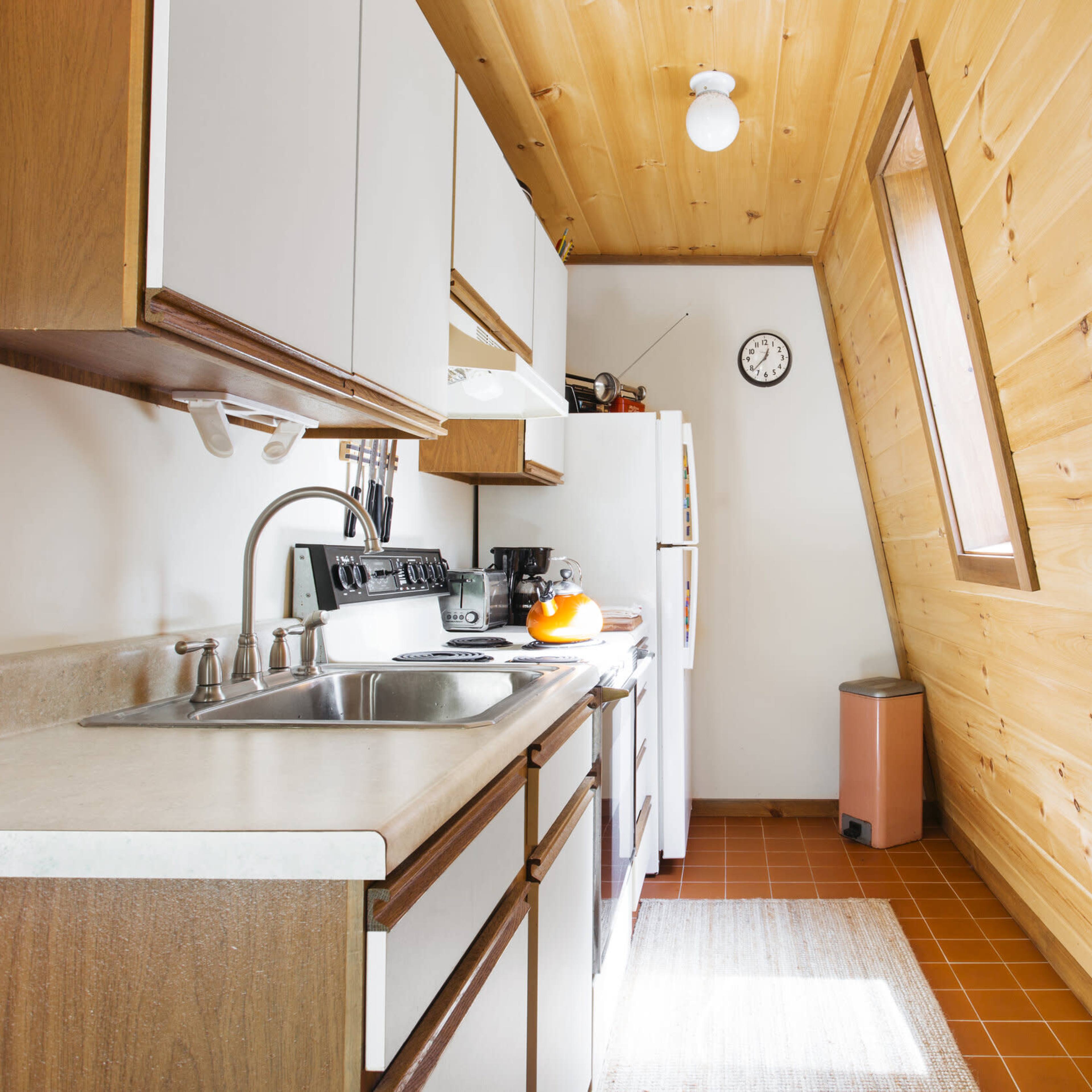 A narrow kitchen with wooden walls, a sink, stove, and white cabinets, featuring a window that tilts open and a clock on the wall.