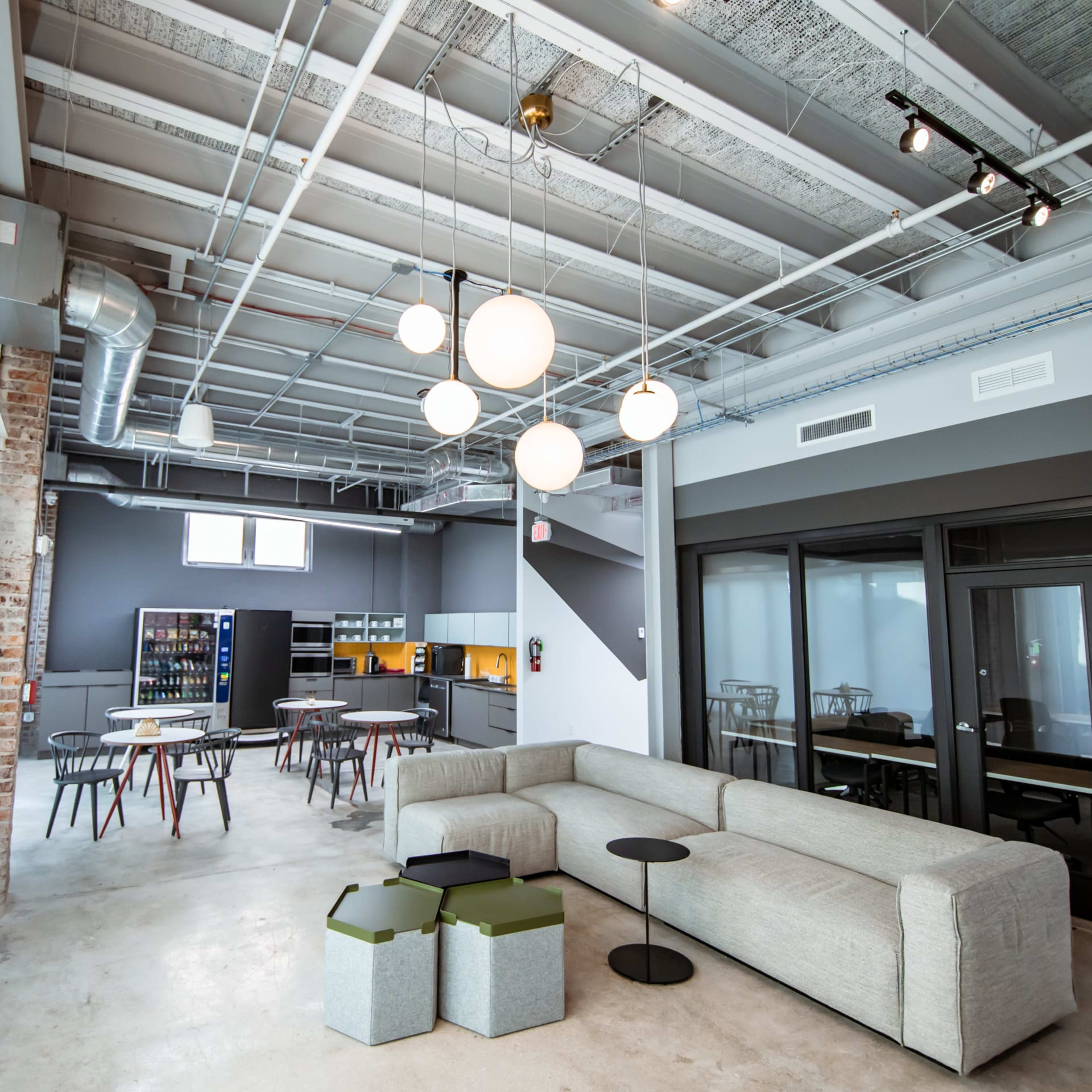 A modern open office space featuring a light-filled area with a grey sectional sofa, round tables, and a kitchenette in the background.