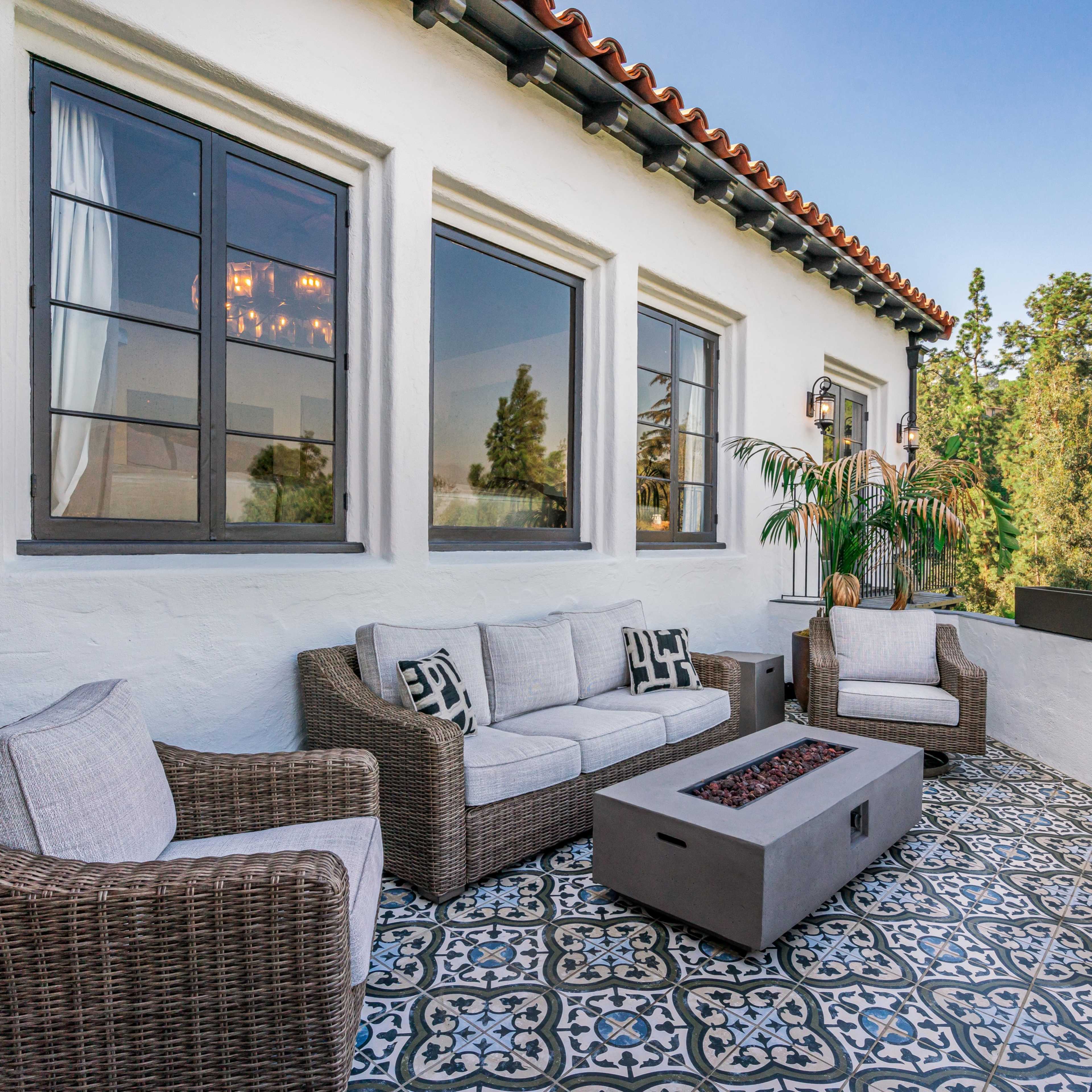 A cozy outdoor patio features a sectional sofa, two chairs, and a coffee table with a fire pit, surrounded by lush greenery.