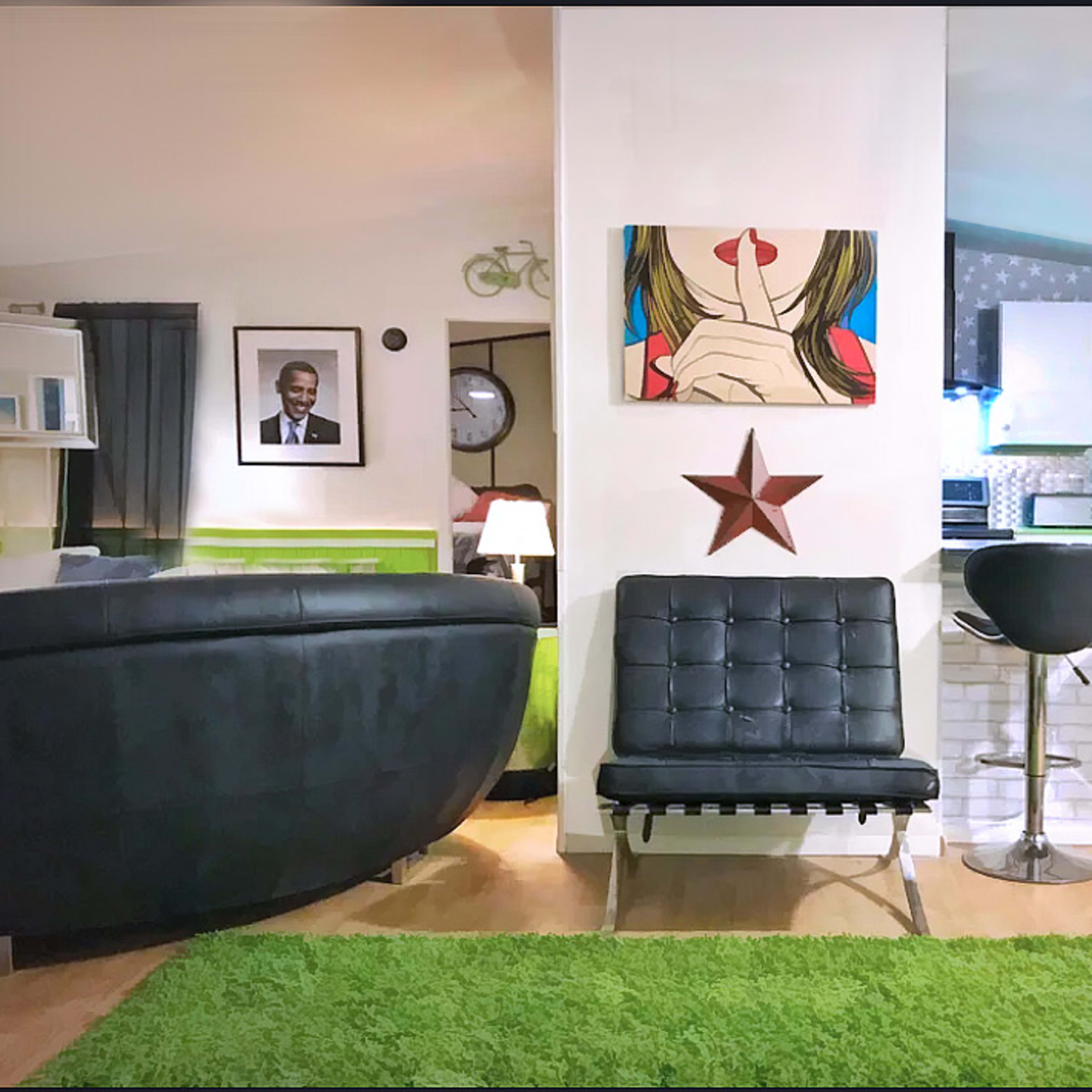 A modern living space featuring a black leather sofa, a green area rug, a star decoration on the wall, and a small bar area with two black stools.