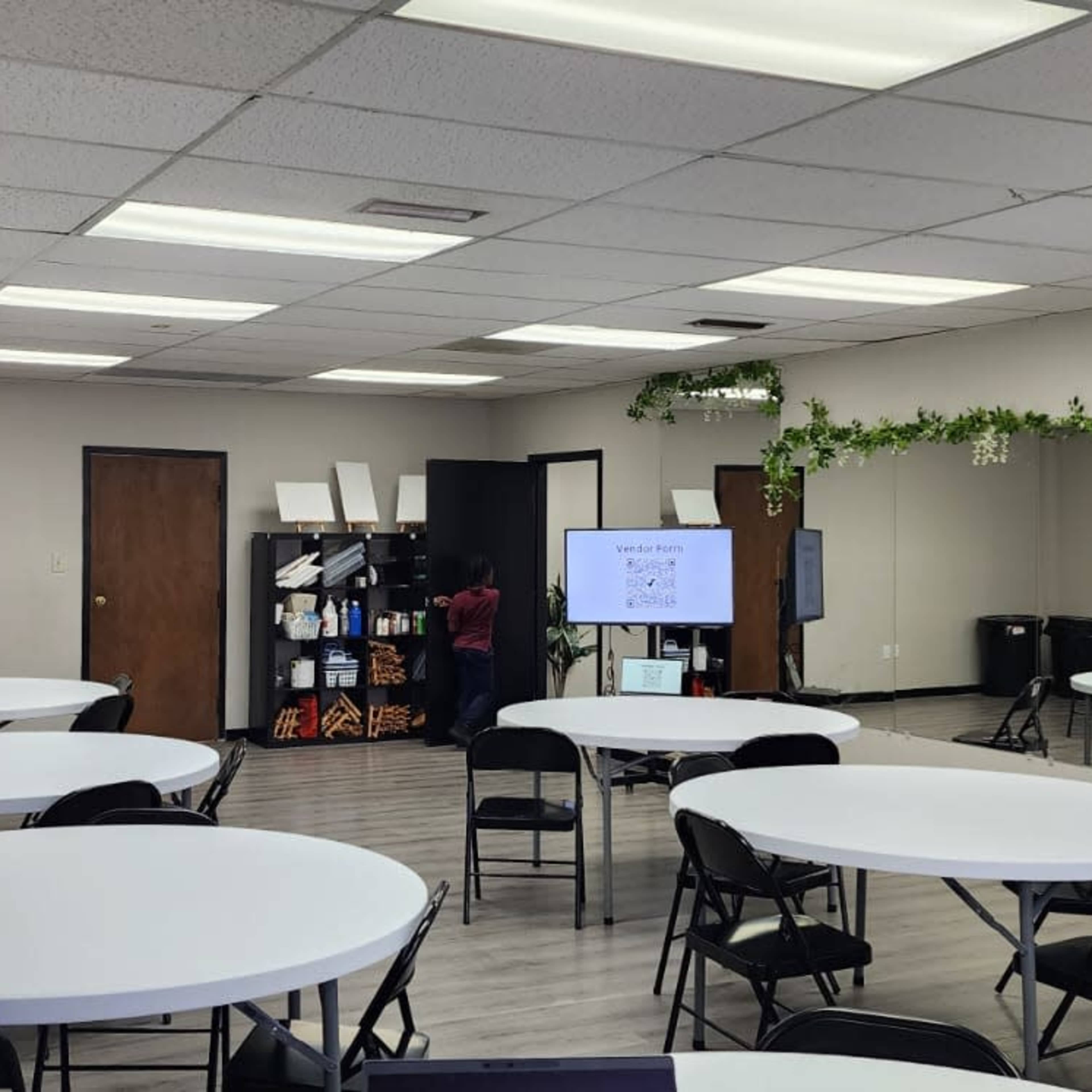 A spacious room with several round tables and black chairs, a large screen, and a mirrored wall, decorated with green plants.