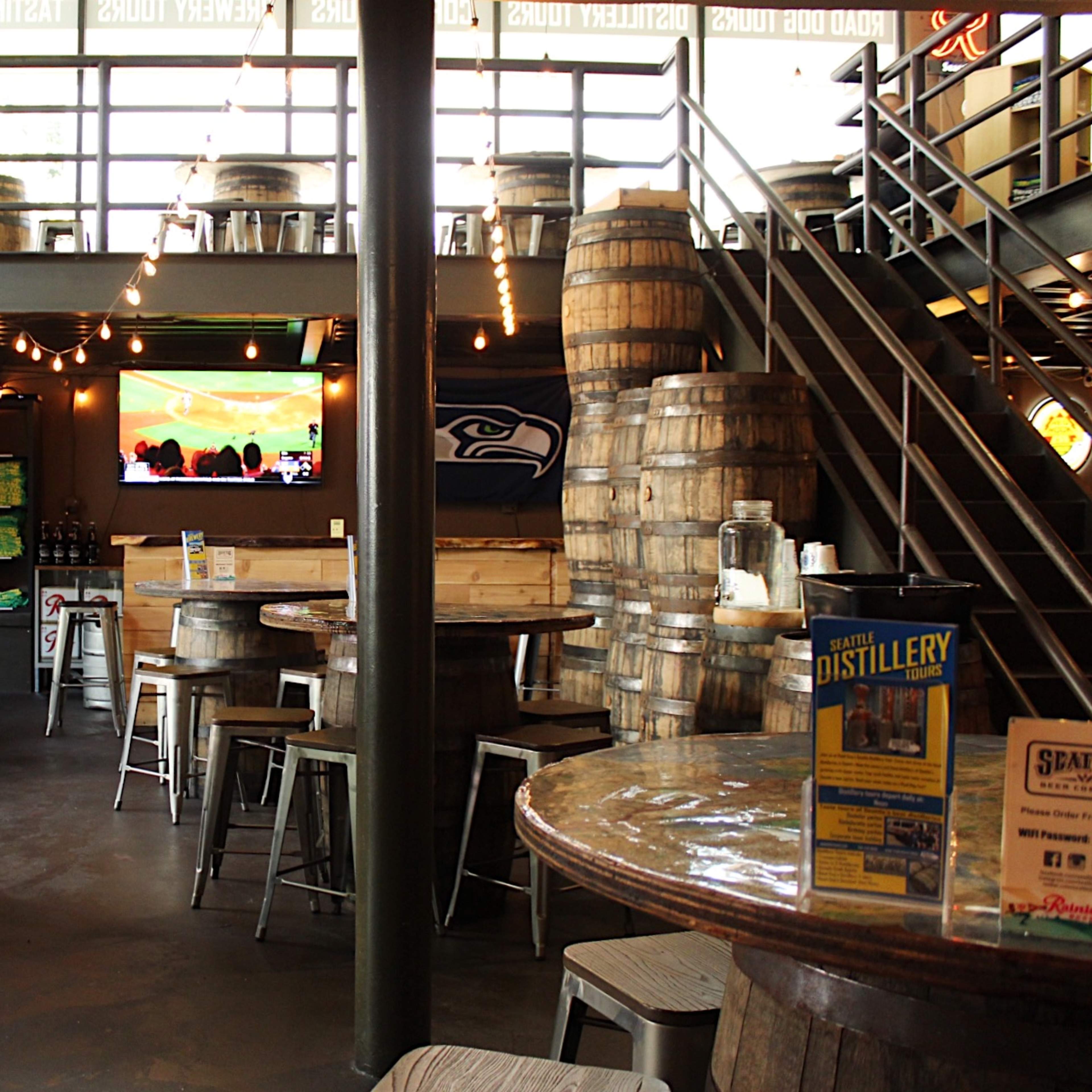 The interior of a brewery features wooden barrel tables and stacks of barrels, with a television displaying a sports event and merchandise on display.