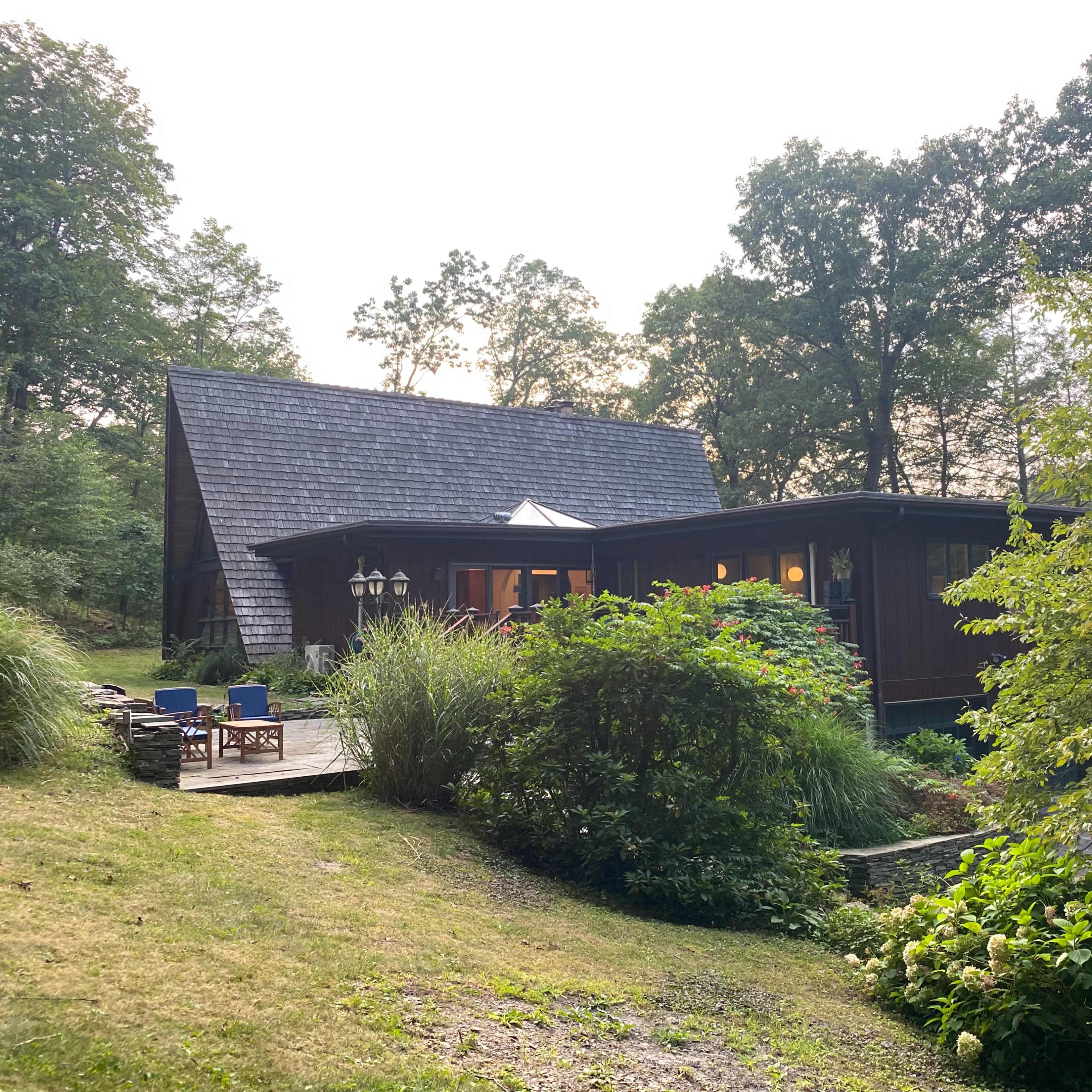 A wooden house with a steep roof sits amid greenery, featuring a porch and large windows that reflect the evening light.