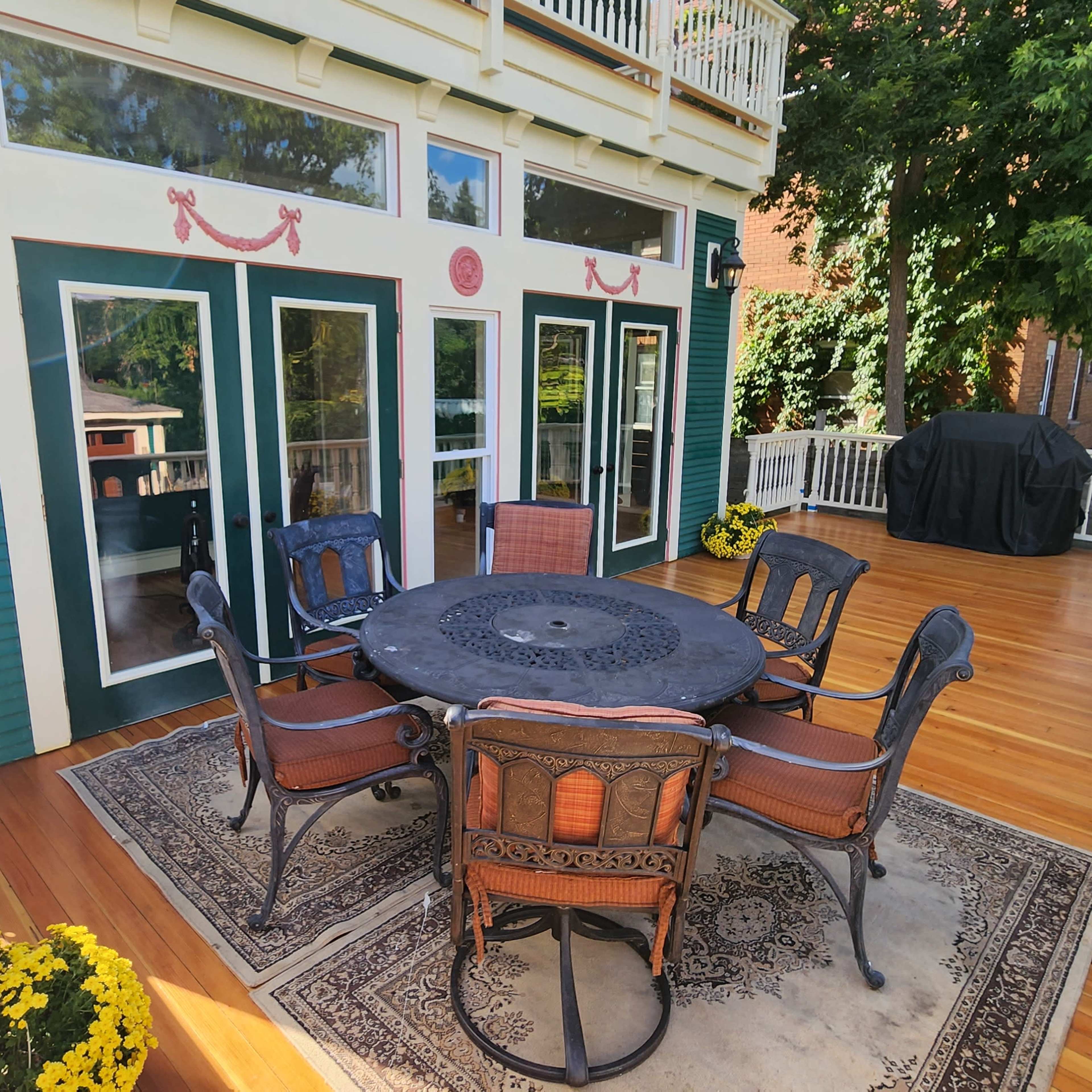 A patio features a round table with six chairs, surrounded by potted yellow flowers and a grill in the background.