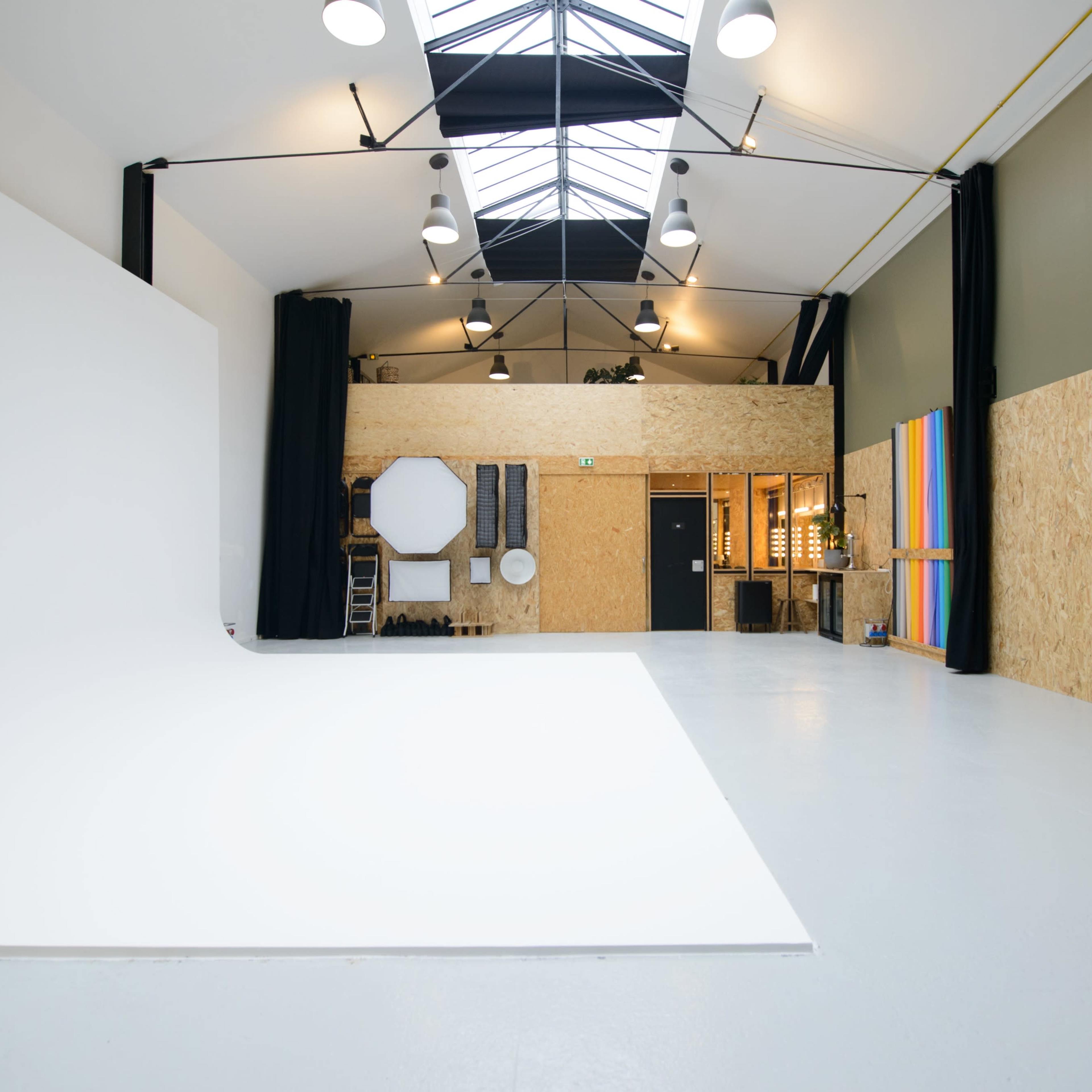 A spacious studio with a blue backdrop on the left, a large white wall, and exposed beams under a skylight.