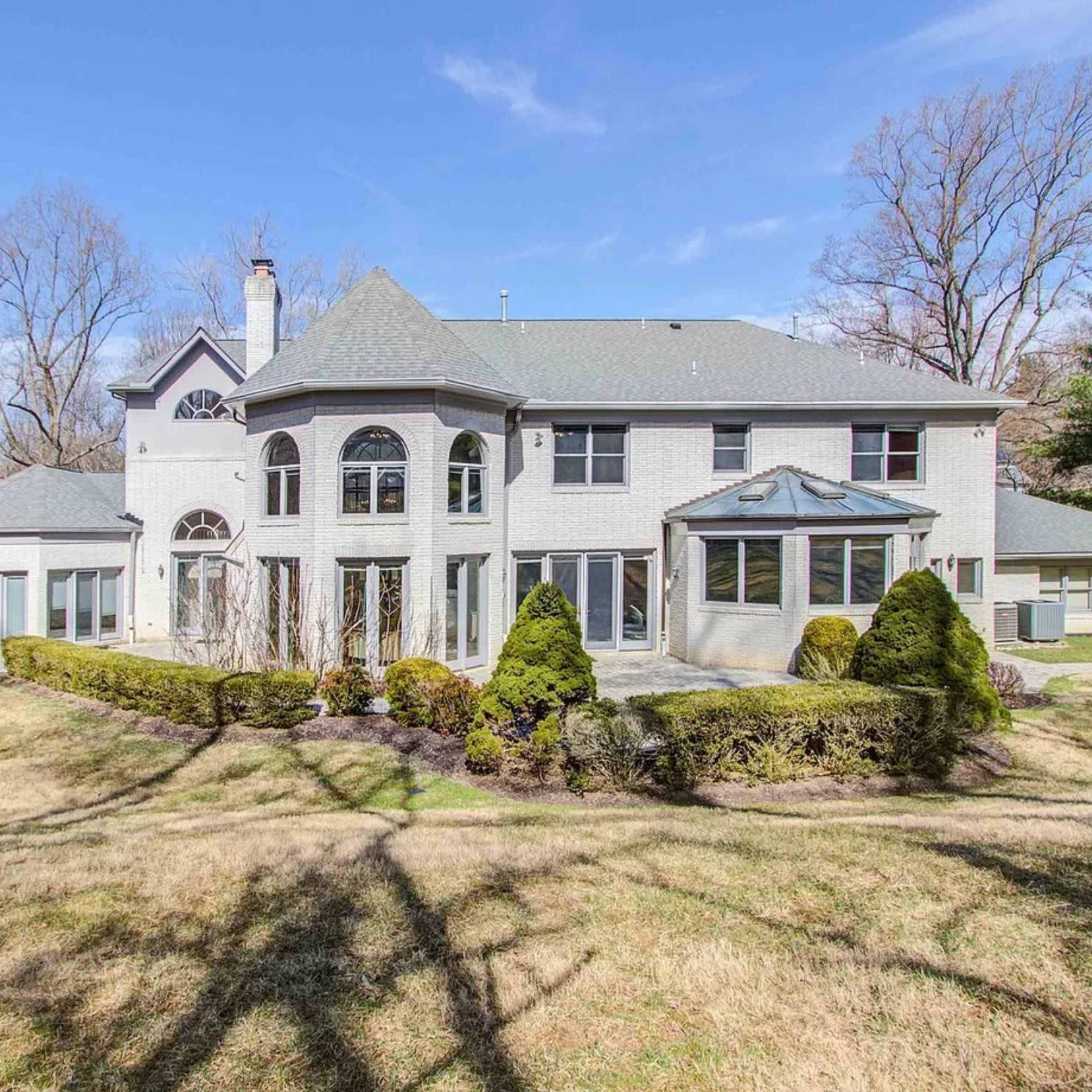 A large, two-story house with multiple gables and windows is set amidst a grassy landscape and bare trees.