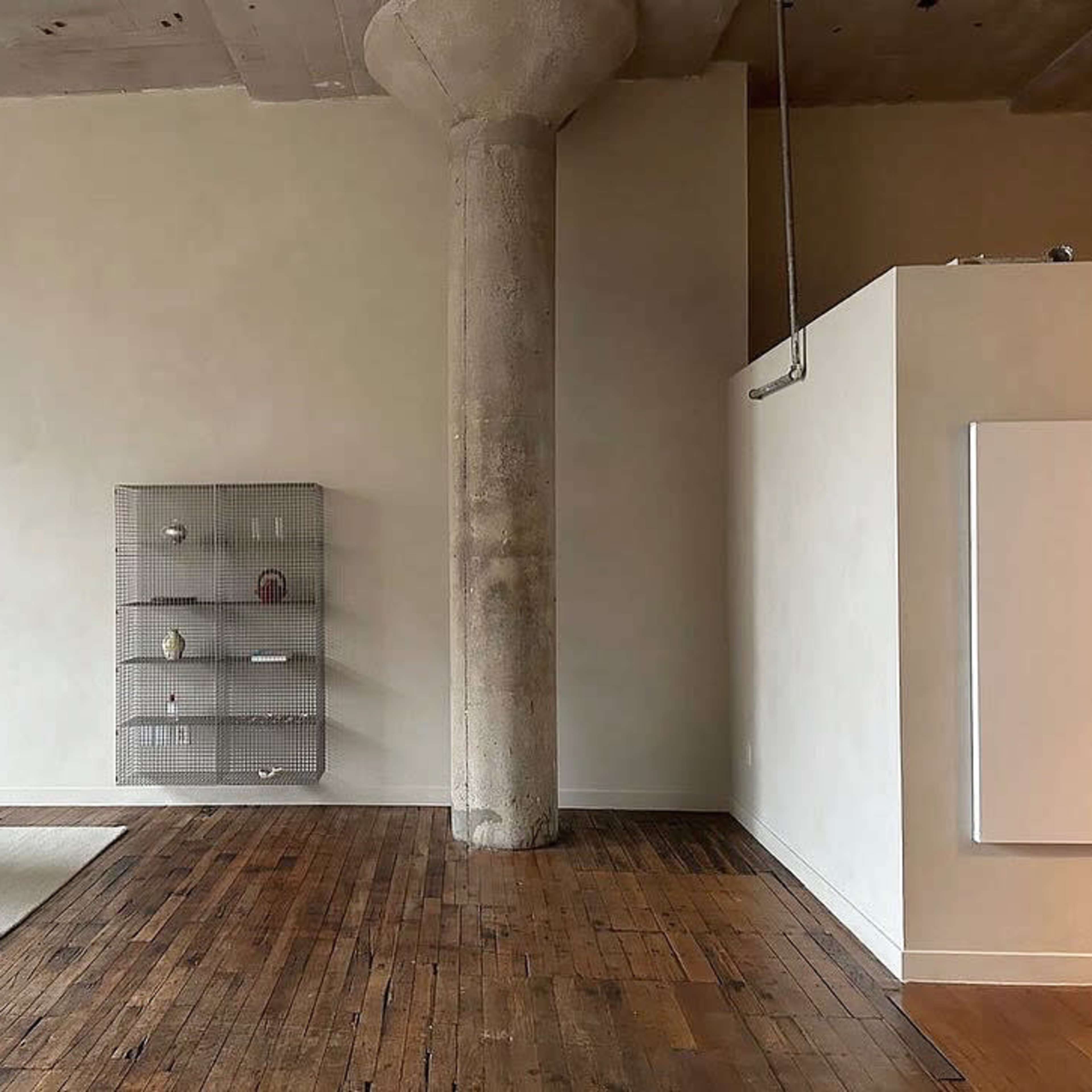 A spacious interior with a wooden floor, a concrete pillar, and a minimalist shelving unit against a light-colored wall.