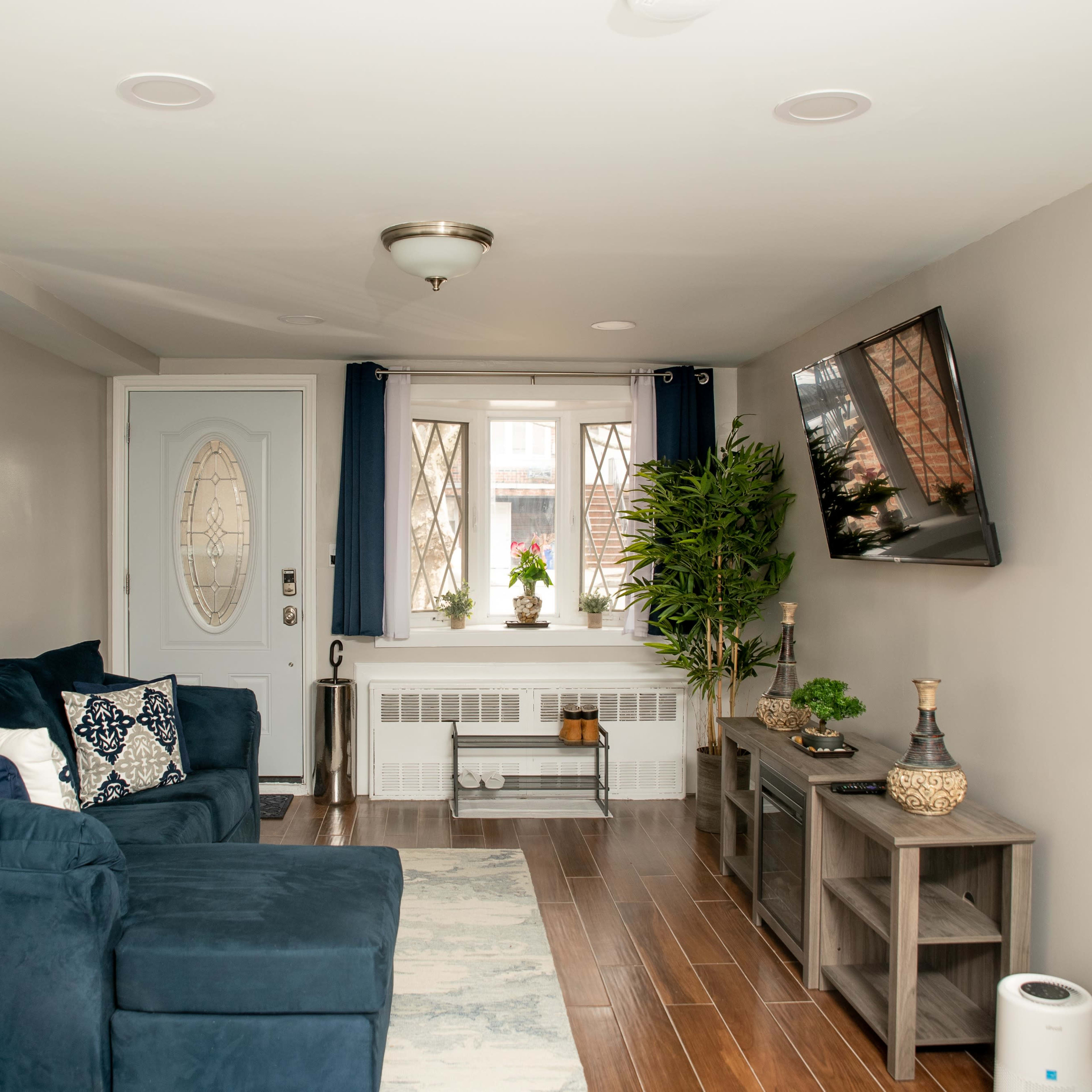 The living room features a blue couch, a TV mounted on the wall, and a window with plants, all arranged around a hardwood floor.