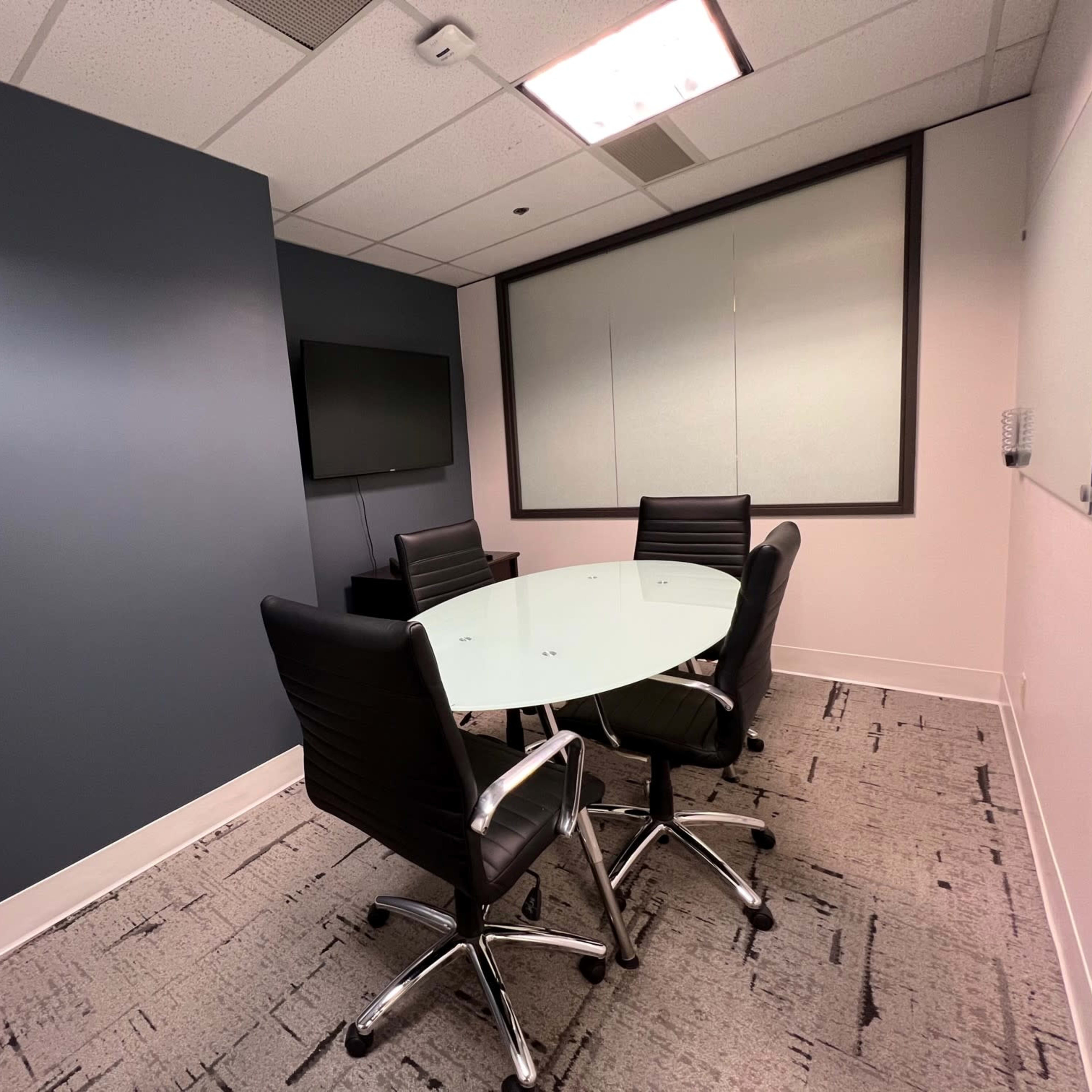 A small conference room features a round glass table surrounded by four black chairs, with a wall-mounted television and a whiteboard.