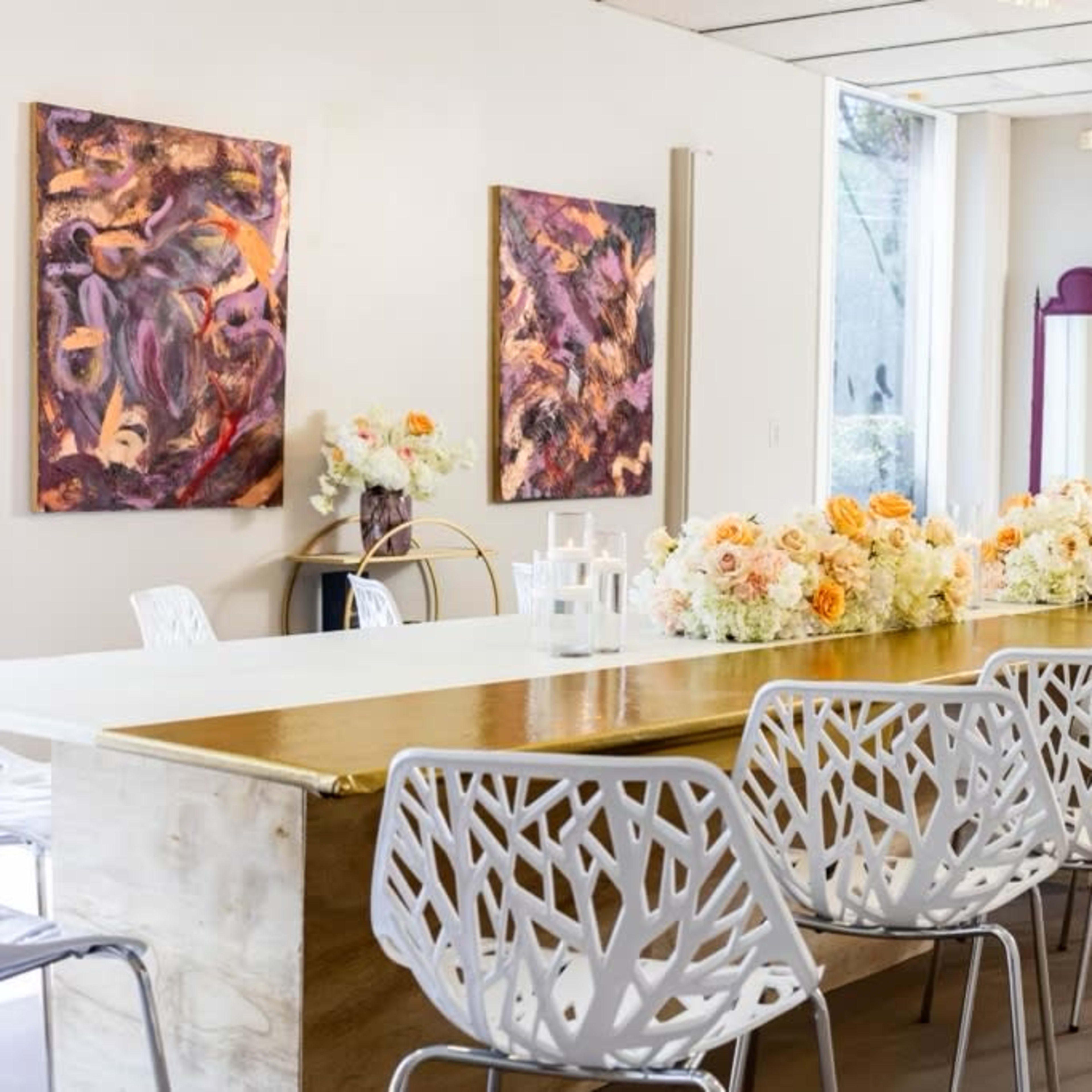 A long wooden table with white, tree-patterned chairs is set up in a well-lit room adorned with floral centerpieces and colorful artwork on the walls.