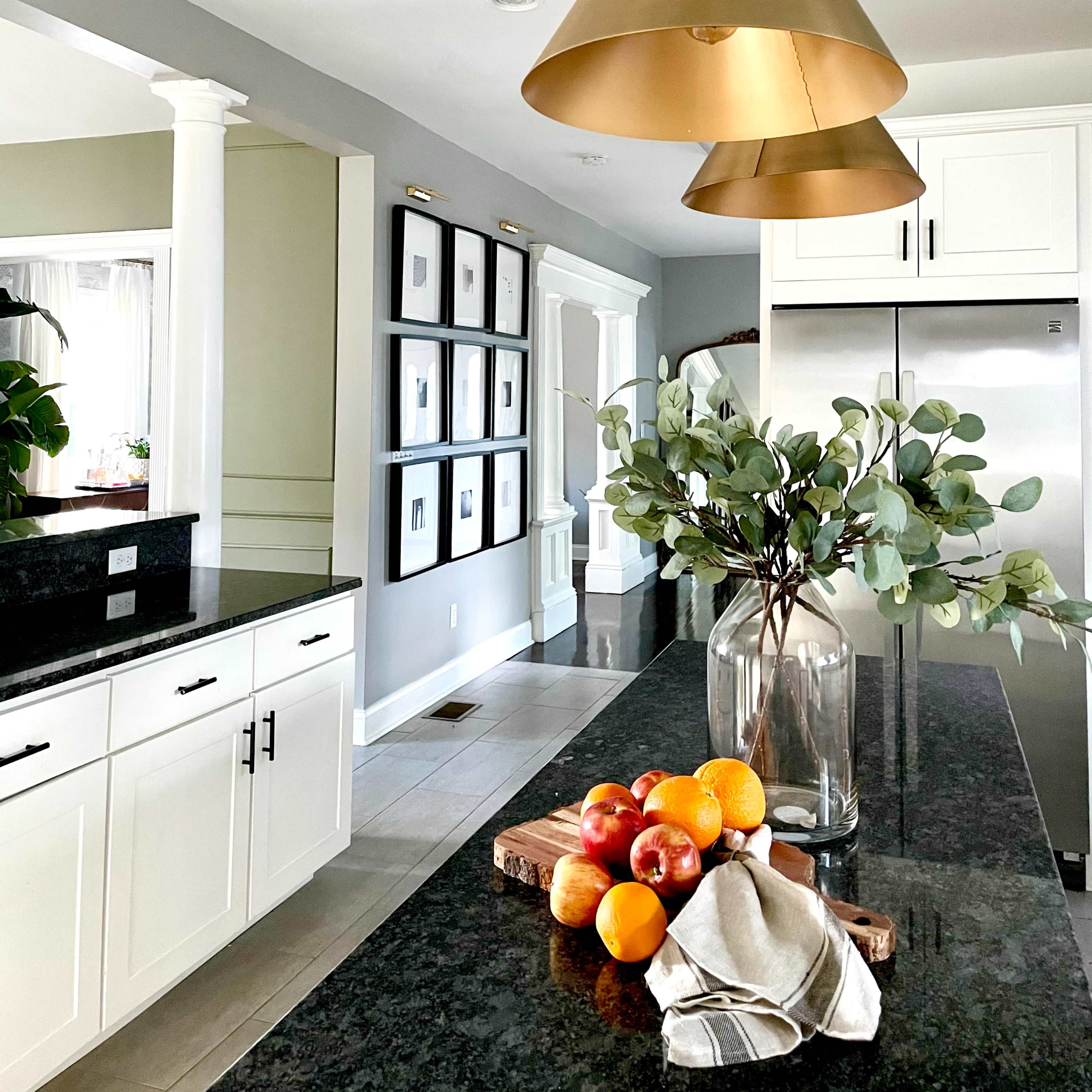 A modern kitchen features brass pendant lights, a black granite countertop with a wooden cutting board holding fruit, and an arrangement of greenery in a glass vase.