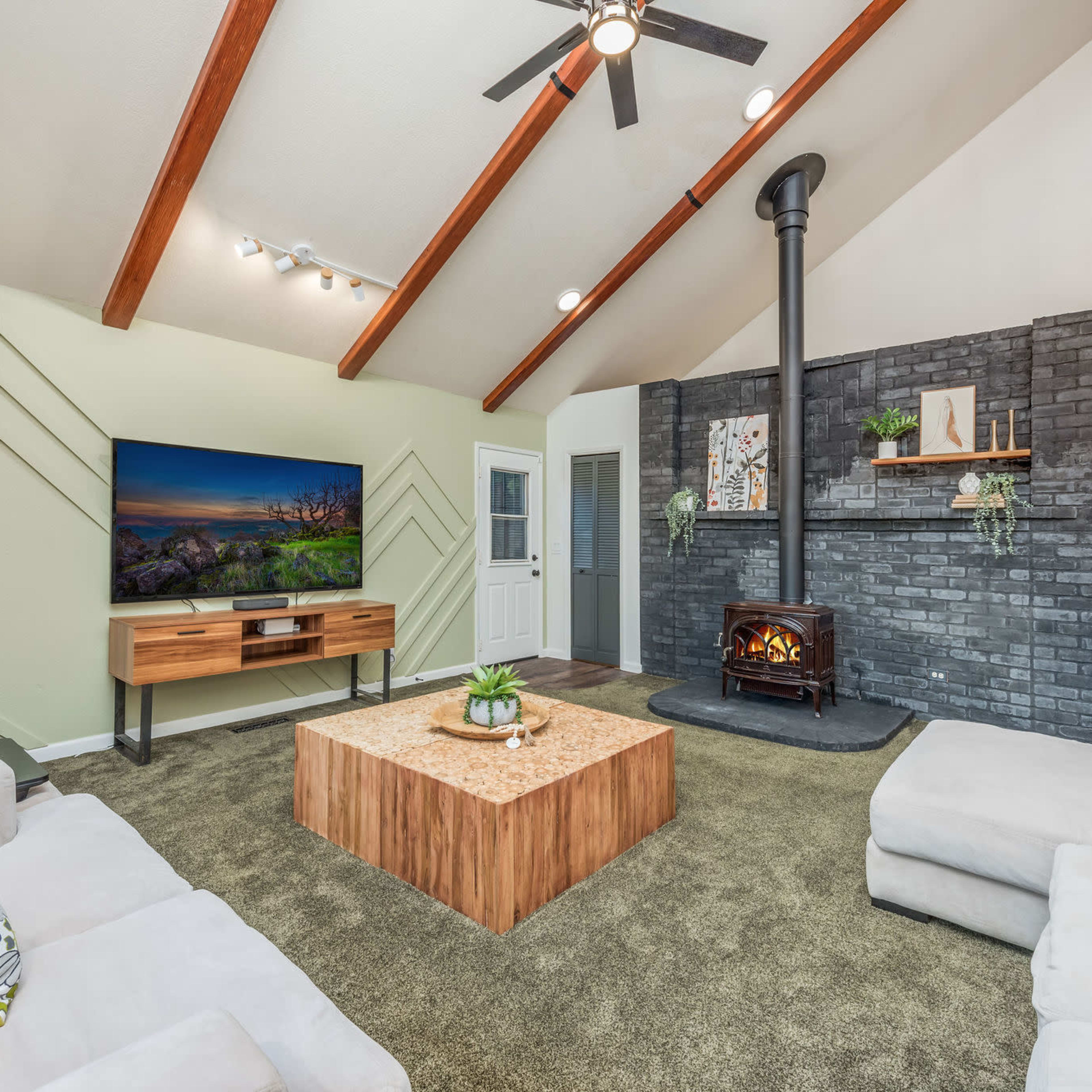 The living room features a wood-burning stove, a large coffee table, and a TV mounted on the wall, with vaulted ceilings and a mix of green and gray decor.