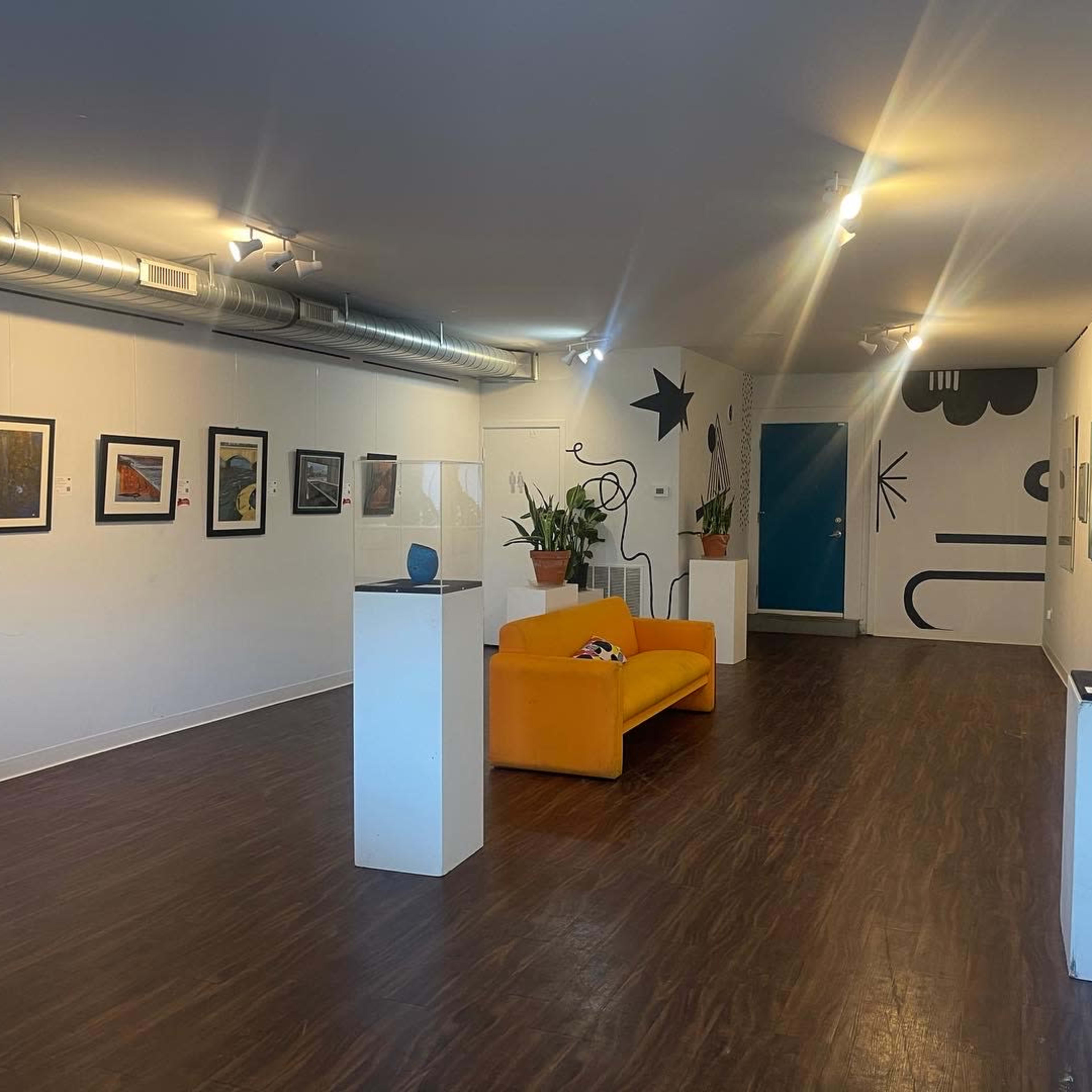 An art gallery with framed artworks on the walls, a bright orange couch in the center, and a few indoor plants near the seating area.