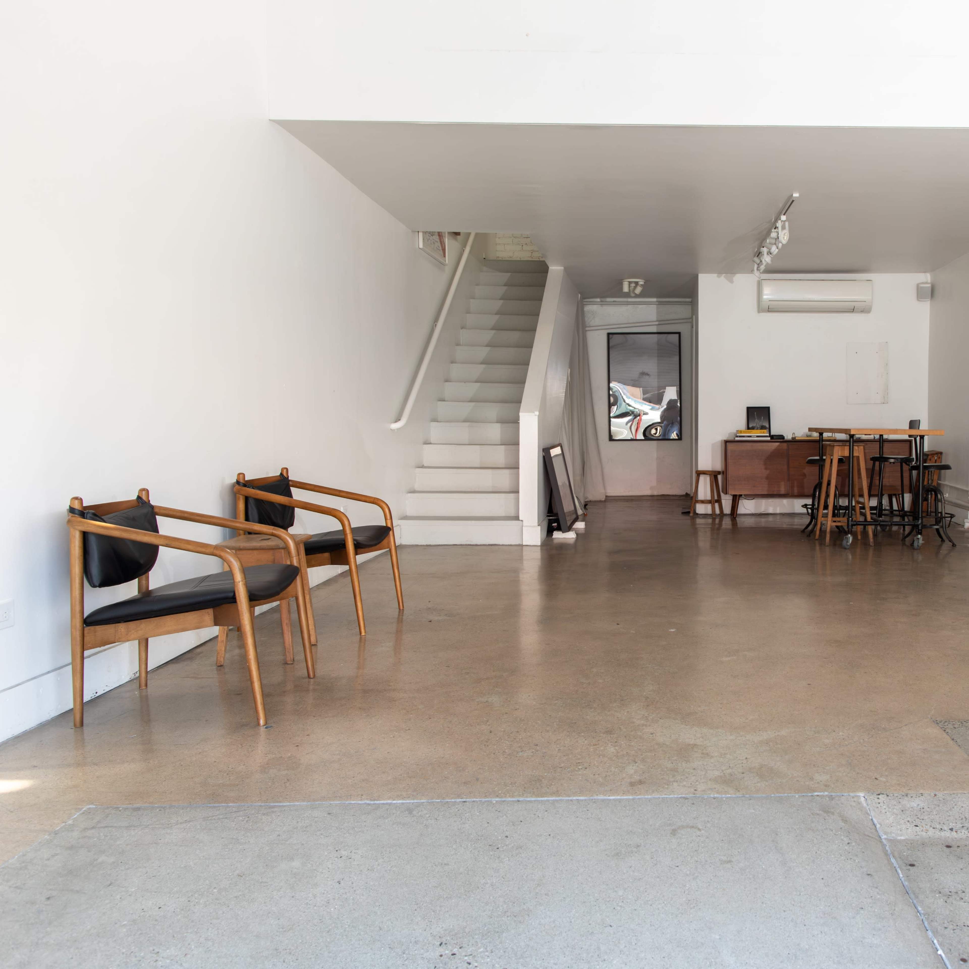 A clean, minimalistic interior space with a staircase, a desk, and a few wooden chairs arranged on polished concrete flooring.