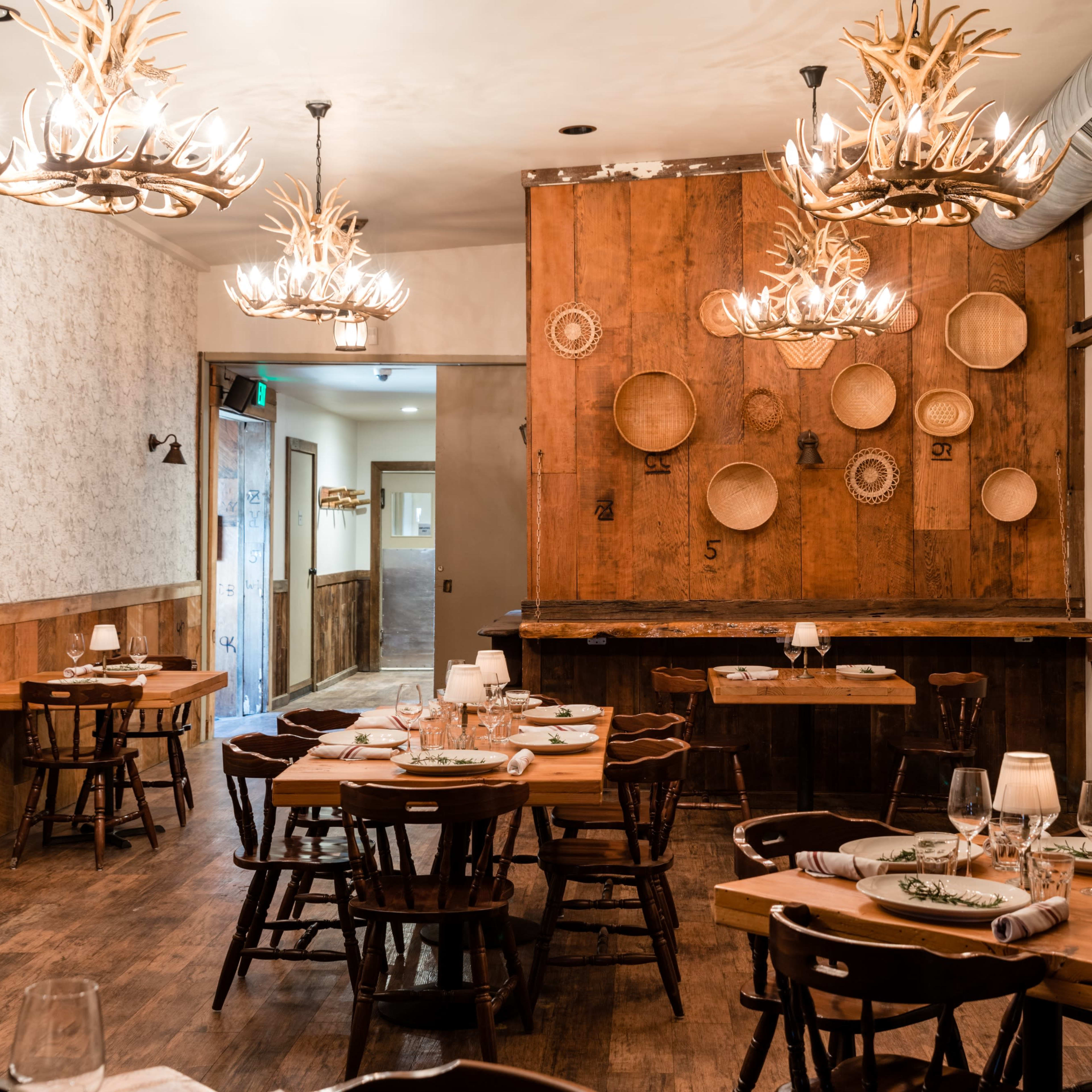A rustic restaurant interior with wooden tables, chairs, and antler chandeliers, featuring wall decorations of woven baskets.
