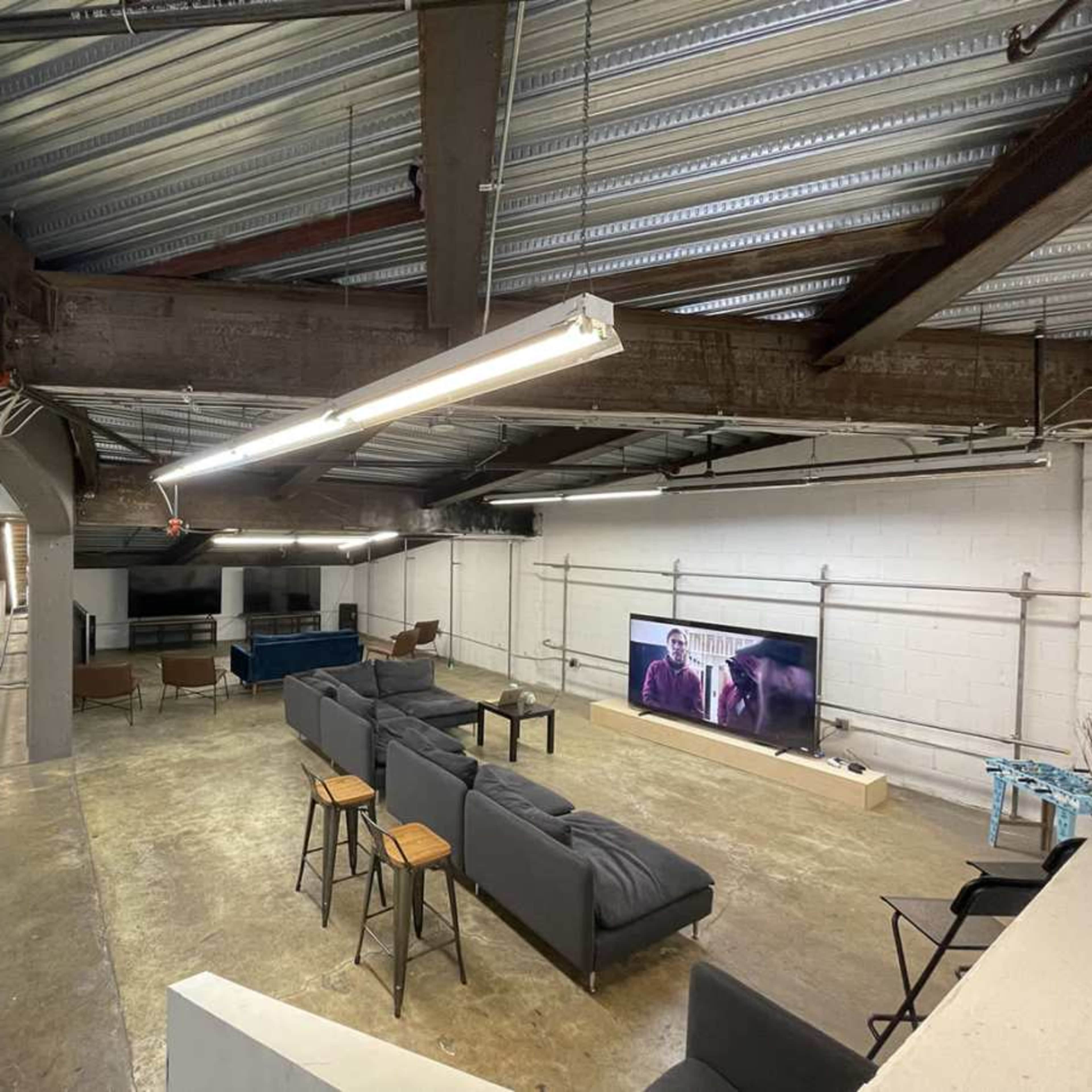 A spacious, industrial-style basement area featuring a seating arrangement with a large sofa and stools, multiple TVs along one wall, and colorful clothing on hangers to the side.
