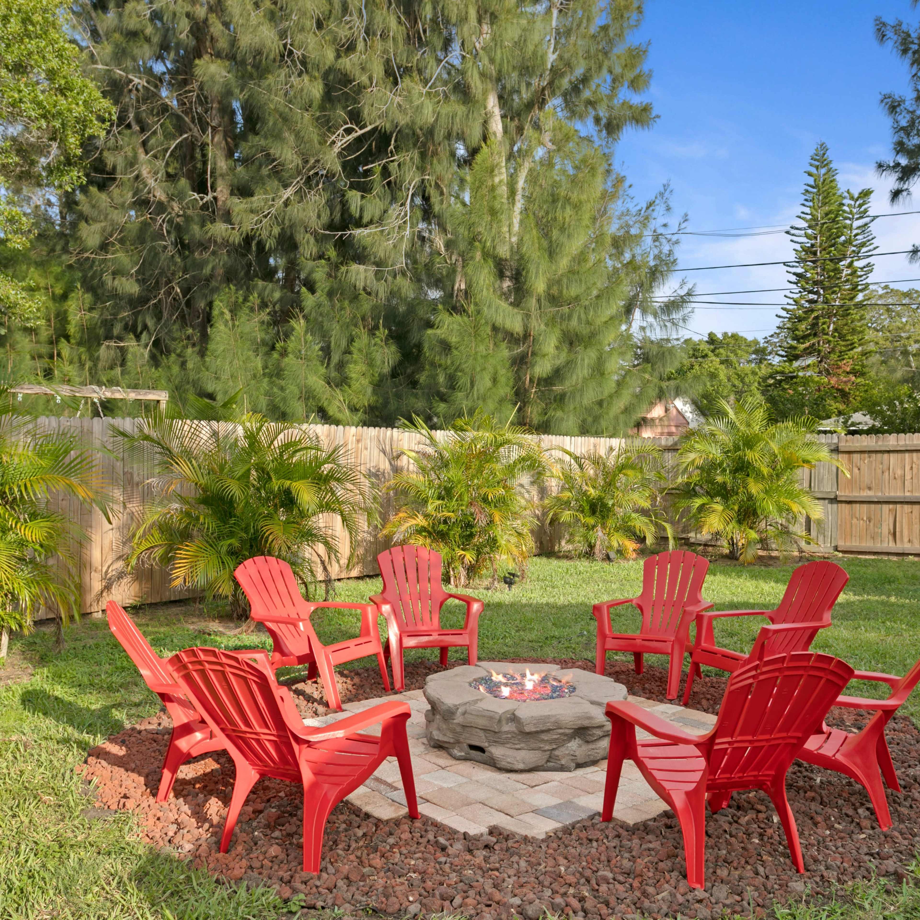 A circular seating arrangement of red chairs surrounds a stone fire pit in a yard with green grass and trees.
