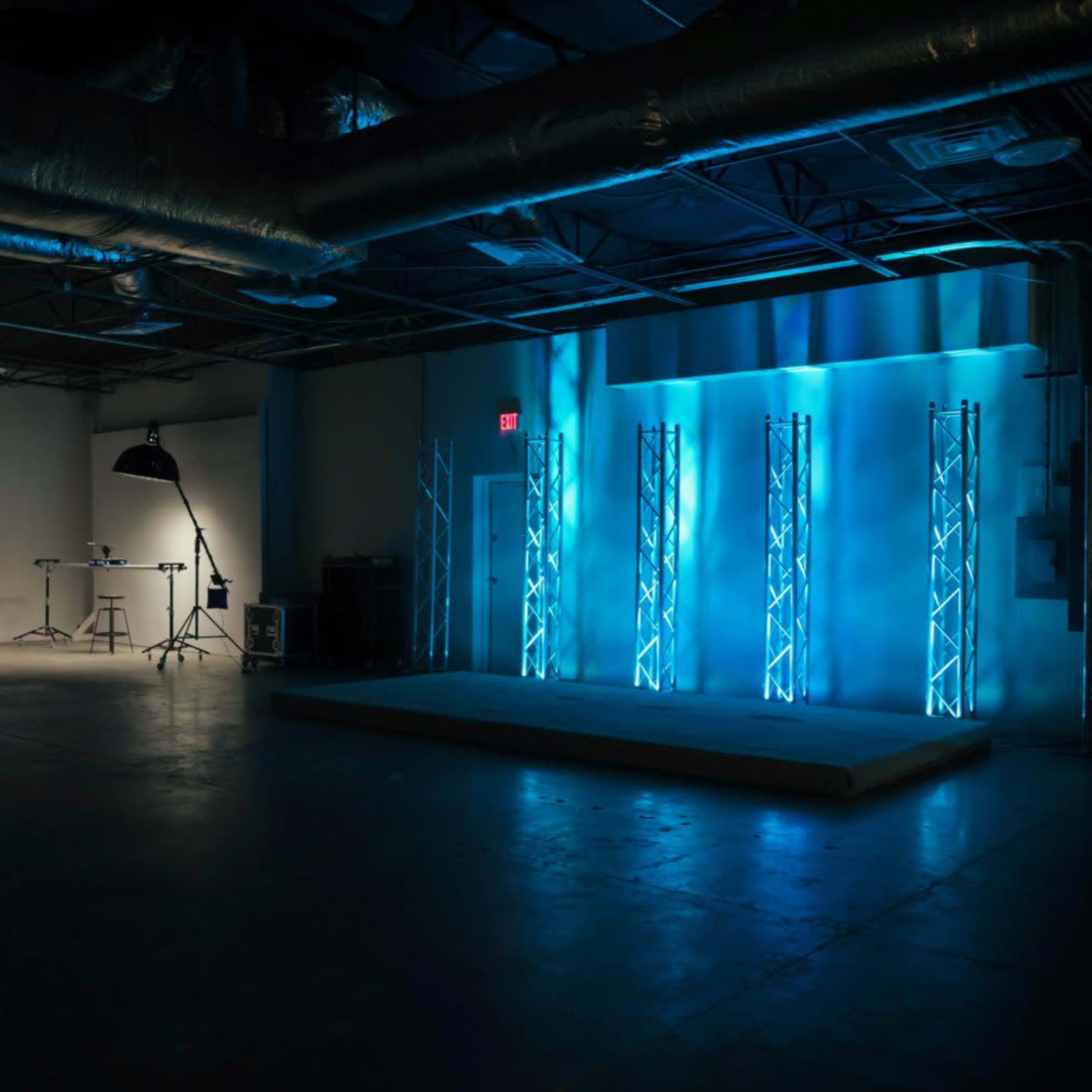A dimly lit studio space featuring a stage with blue lighting, a row of empty chairs, and various lighting equipment set up.