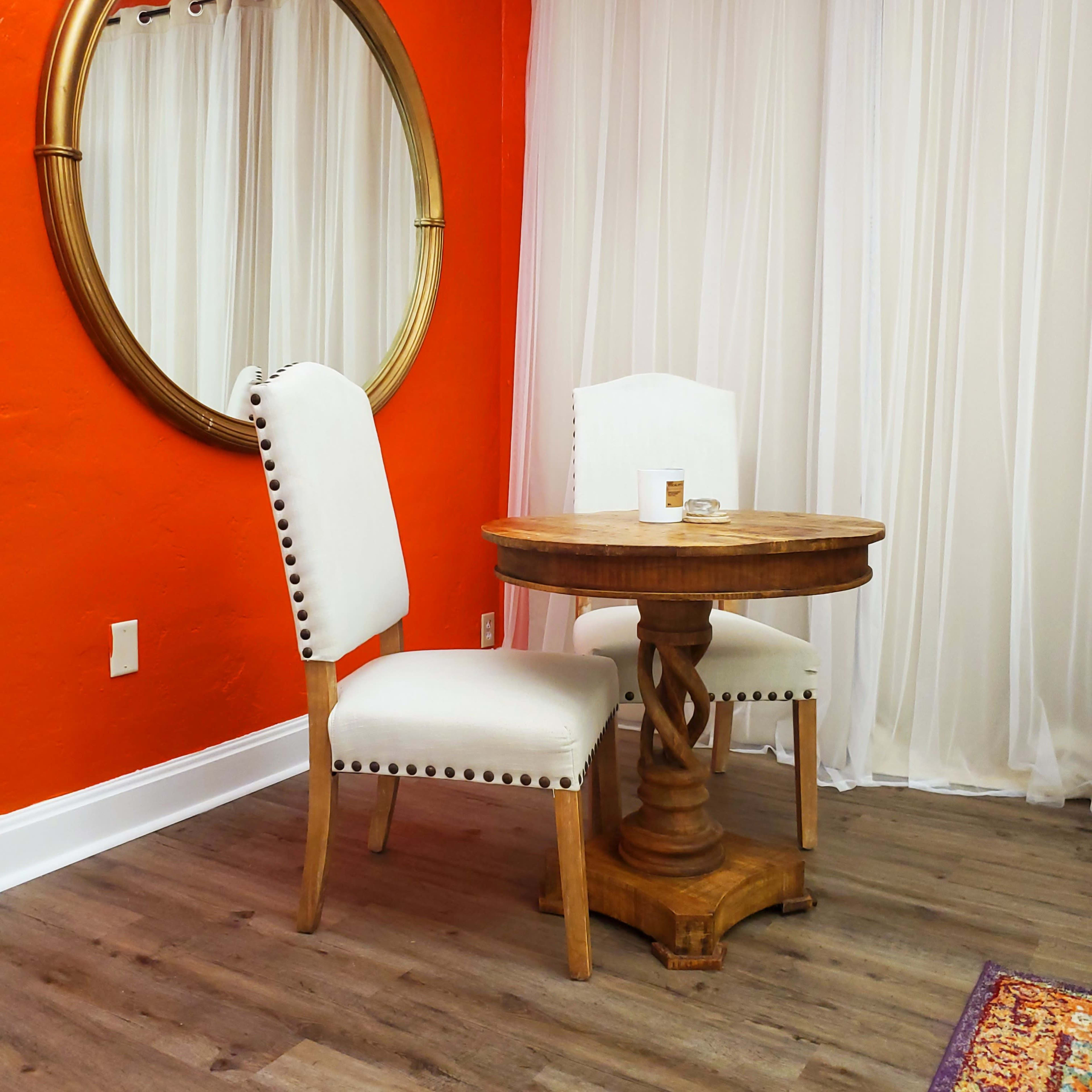 A round wooden table with two upholstered chairs is set against an orange wall, with a large round mirror hanging above.