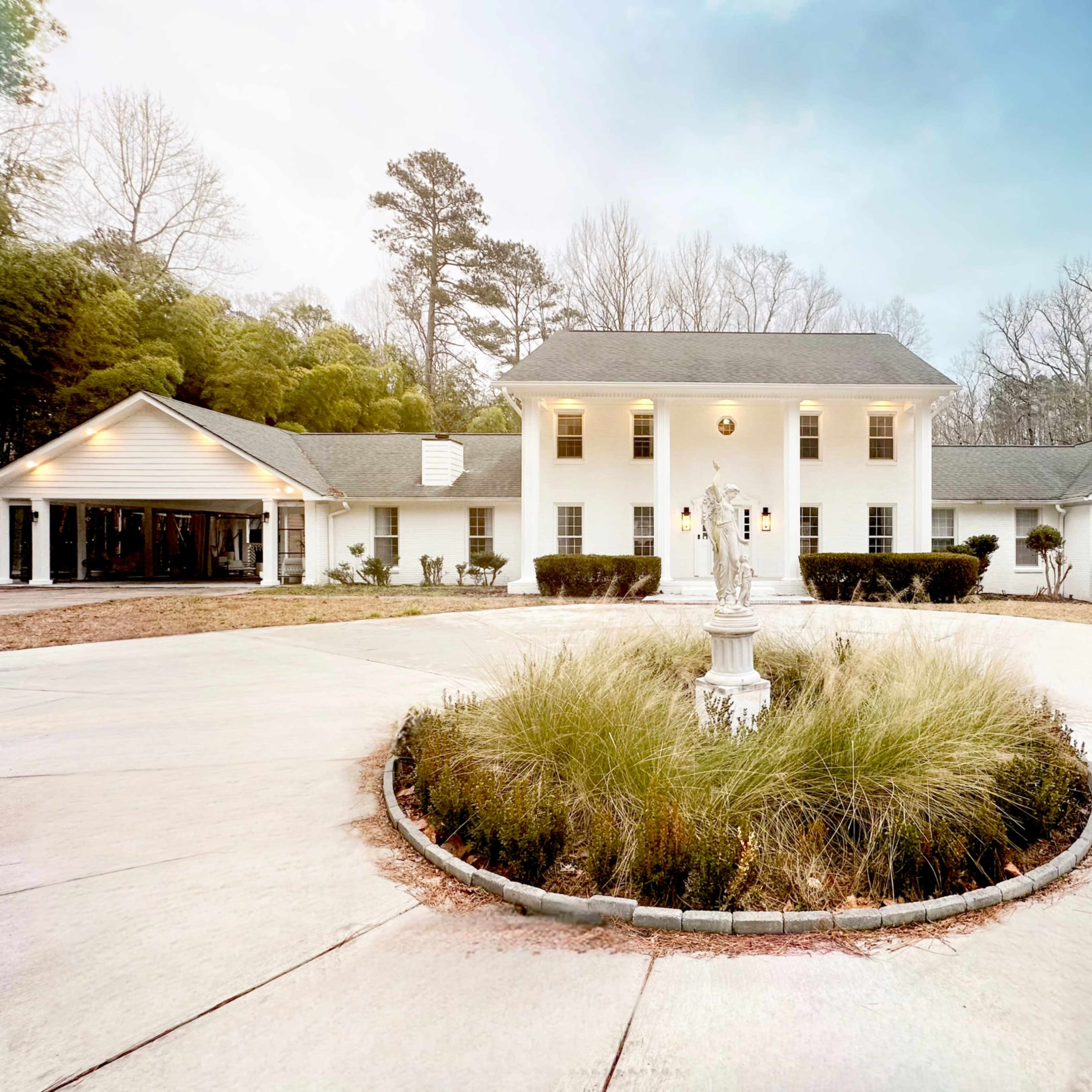 A large white house with a circular driveway and a central fountain is surrounded by neatly trimmed shrubs and trees.