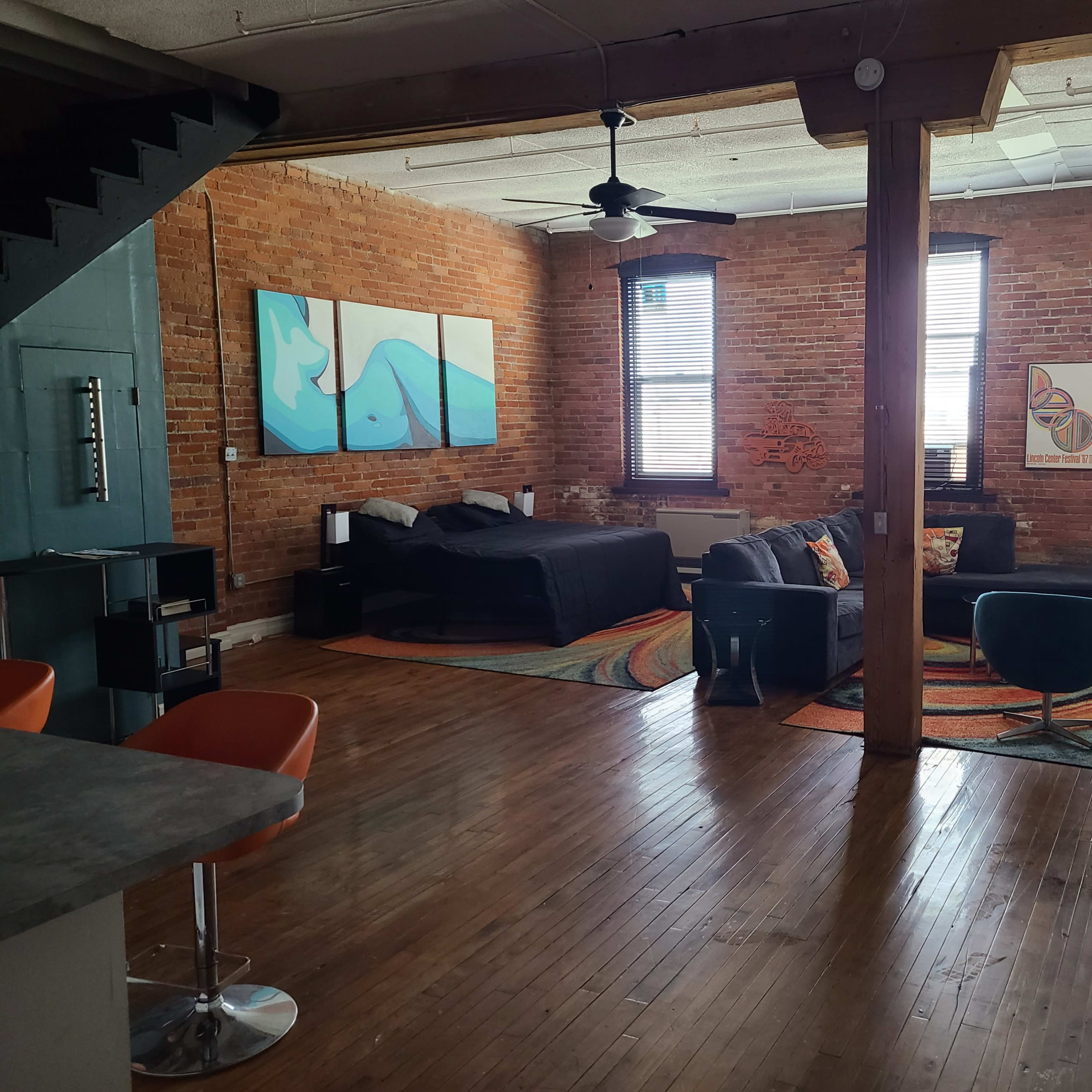 A spacious loft-style living area with exposed brick walls, large windows, a black bed, and modern furniture arranged on a colorful patterned rug.