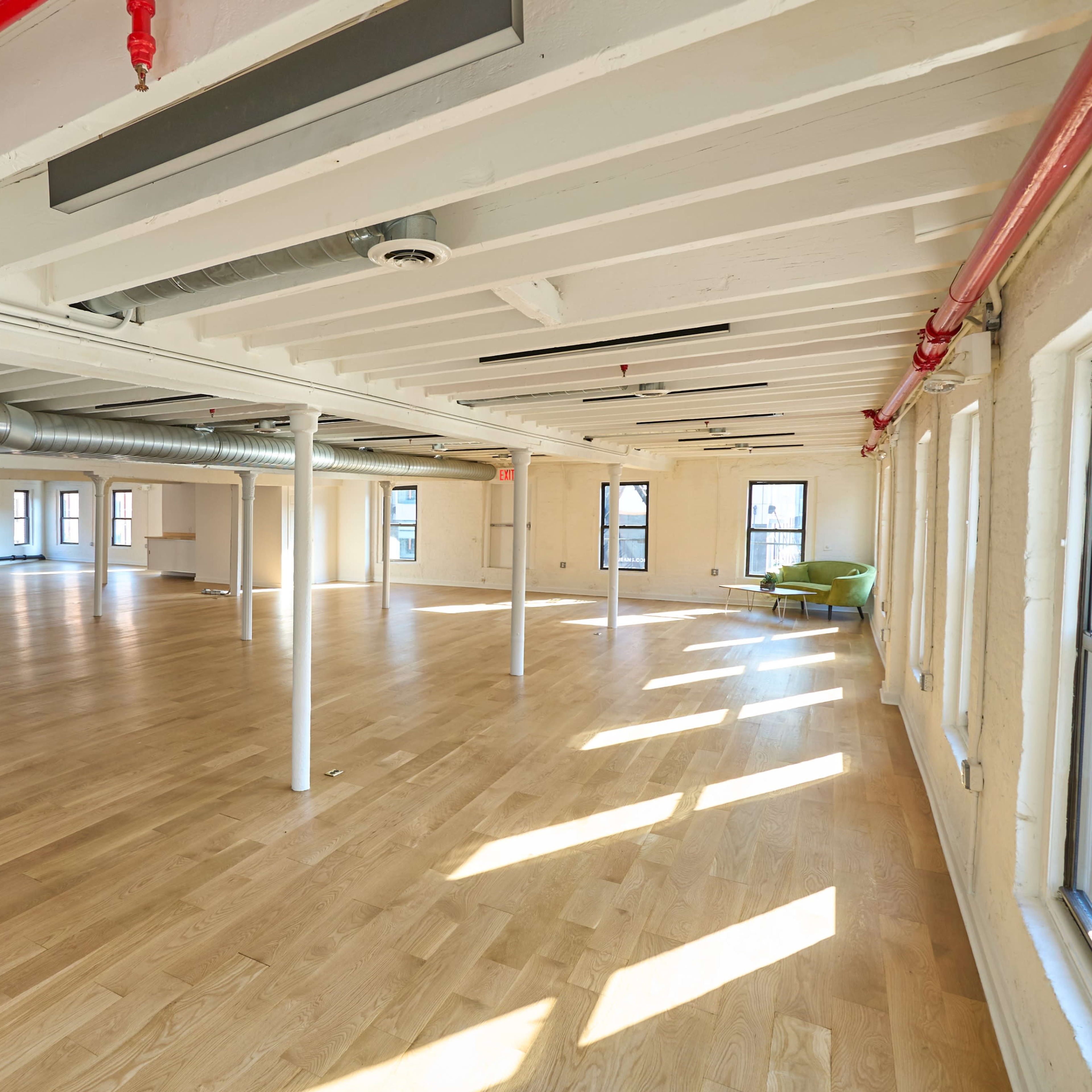 A spacious, empty interior room with wooden flooring, exposed beams, and large windows allowing natural light to illuminate the space.