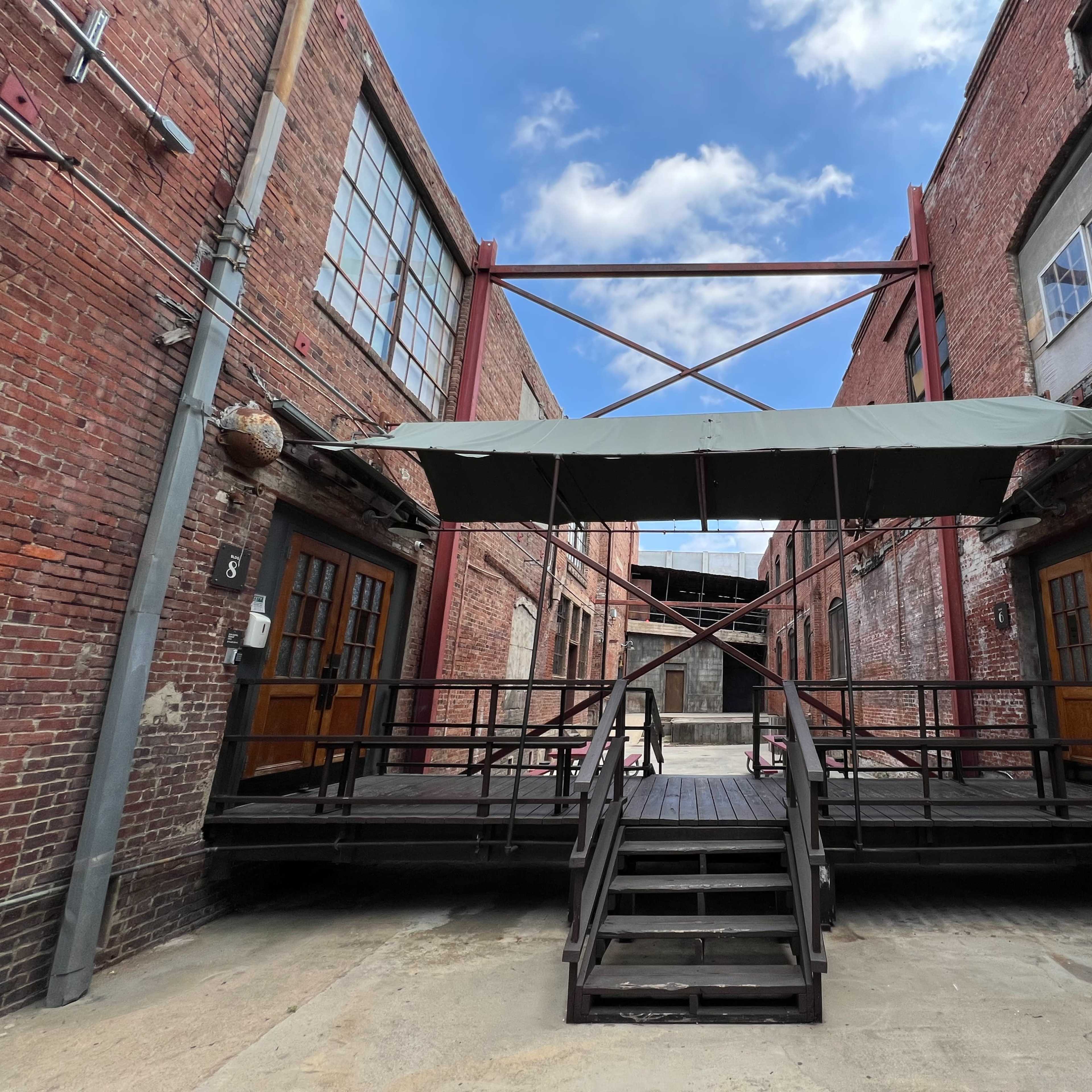 A brick building complex with a wooden deck and a large canopy supported by metal beams, leading to two double doors.
