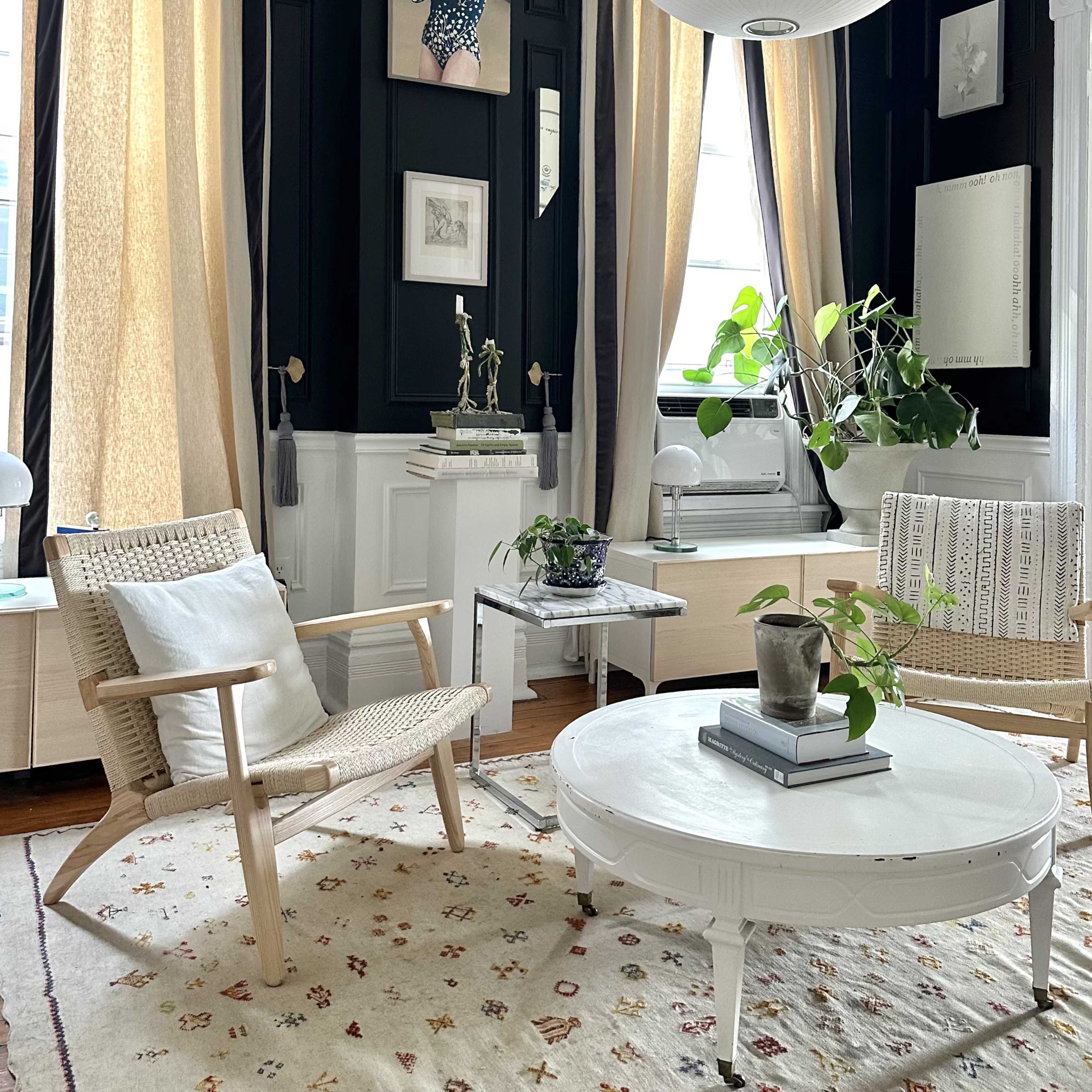 A living room features a round coffee table, two woven chairs, a patterned rug, and tall curtains, with art displayed on the walls and a potted plant in the space.