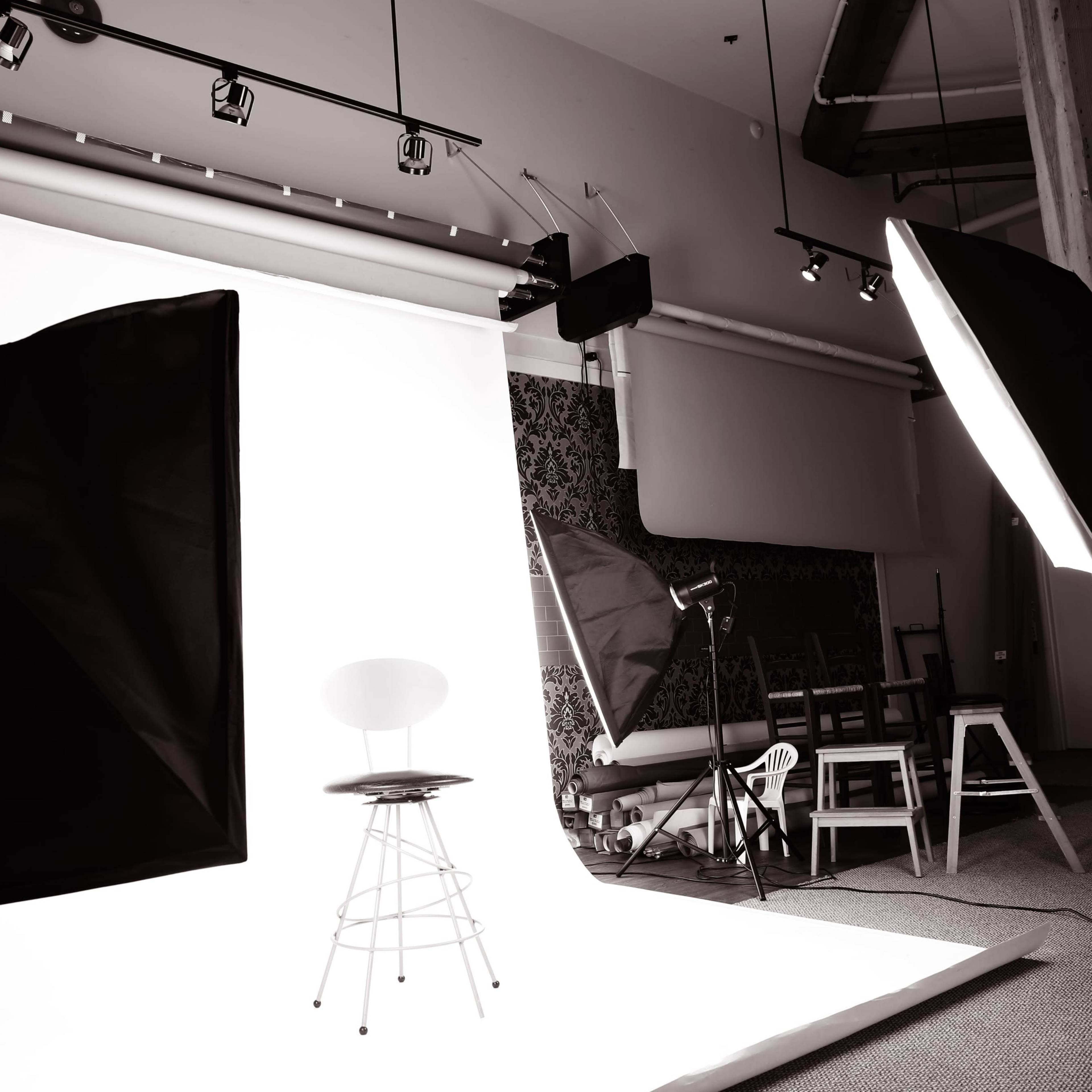 A photography studio features a simple backdrop with lighting equipment and an empty stool positioned for a photoshoot.