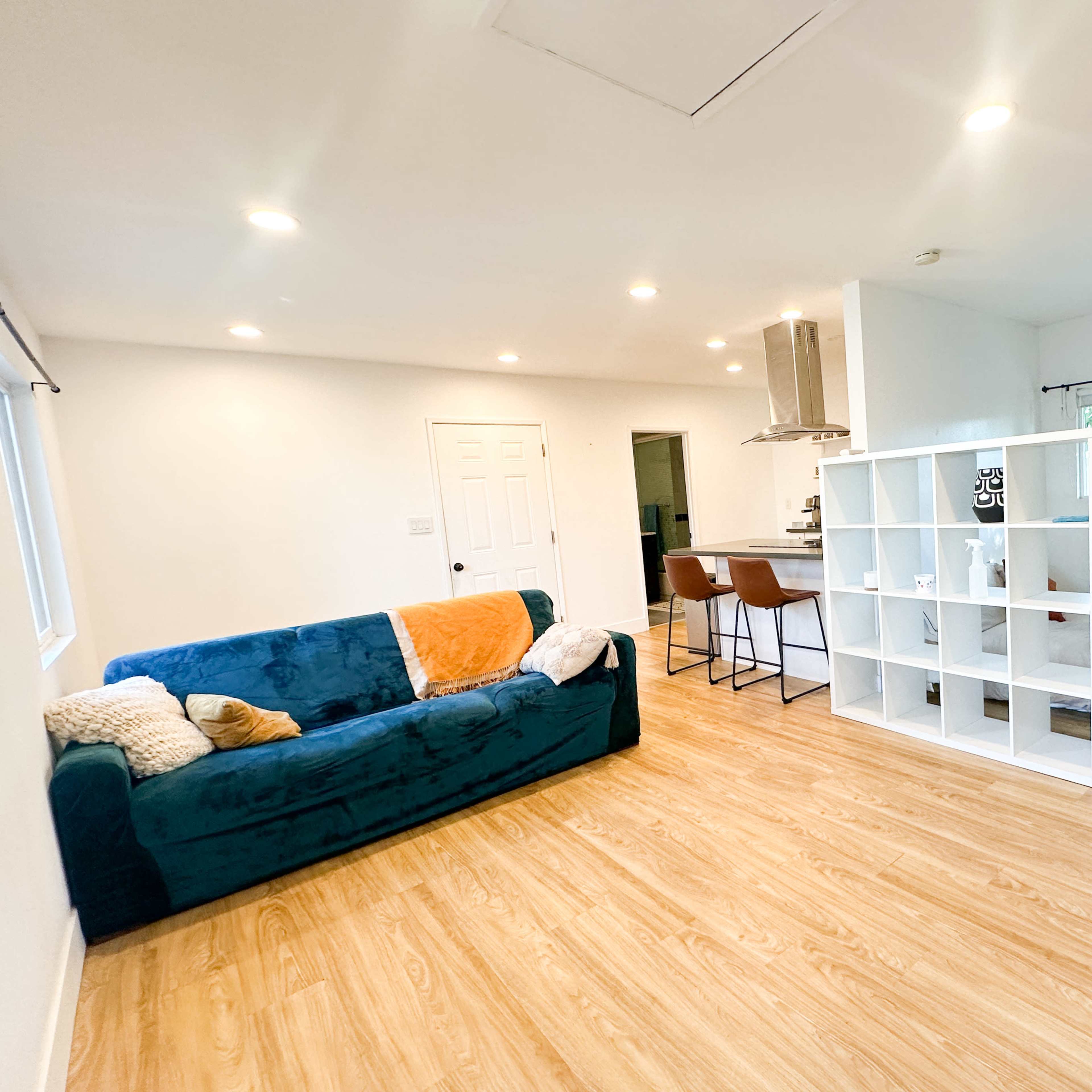 A modern living space with a teal couch, a shelving unit, and a kitchen area with bar seating.