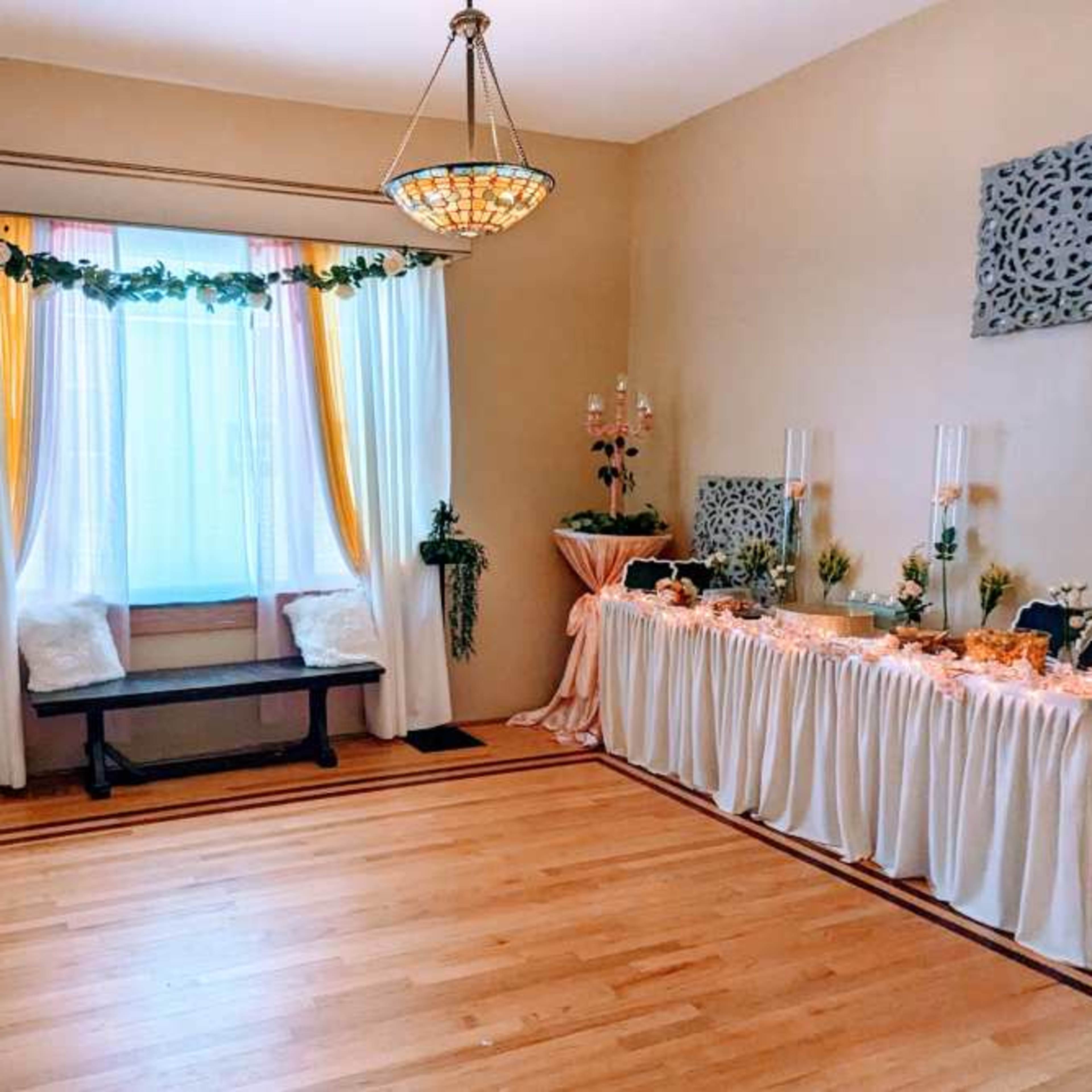The room features a window adorned with curtains, a wooden bench beneath it, and a decorative table set with tablecloths and various floral arrangements.