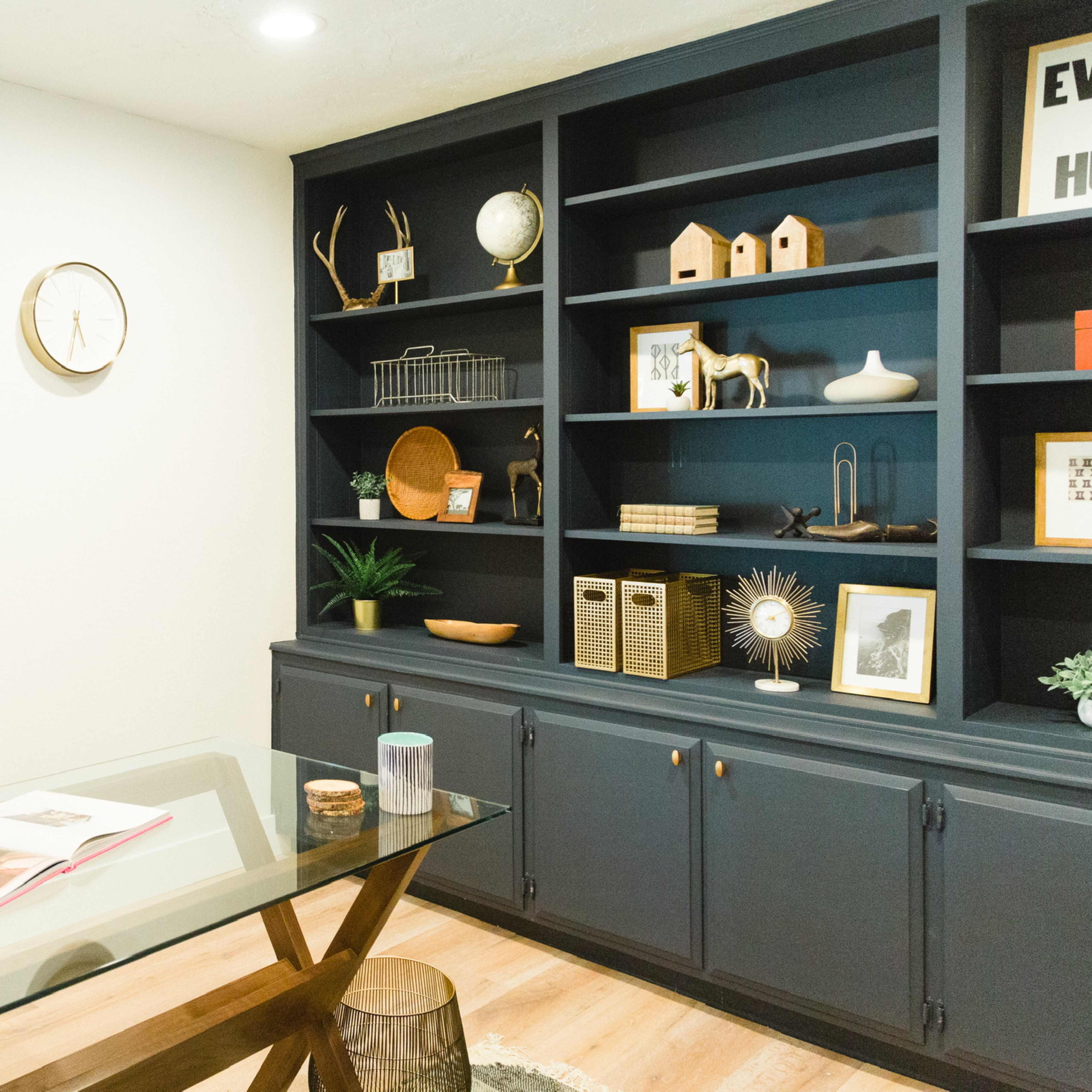 A modern office space with a glass desk, a black chair, and a stylish bookshelf filled with decorative items and plants.