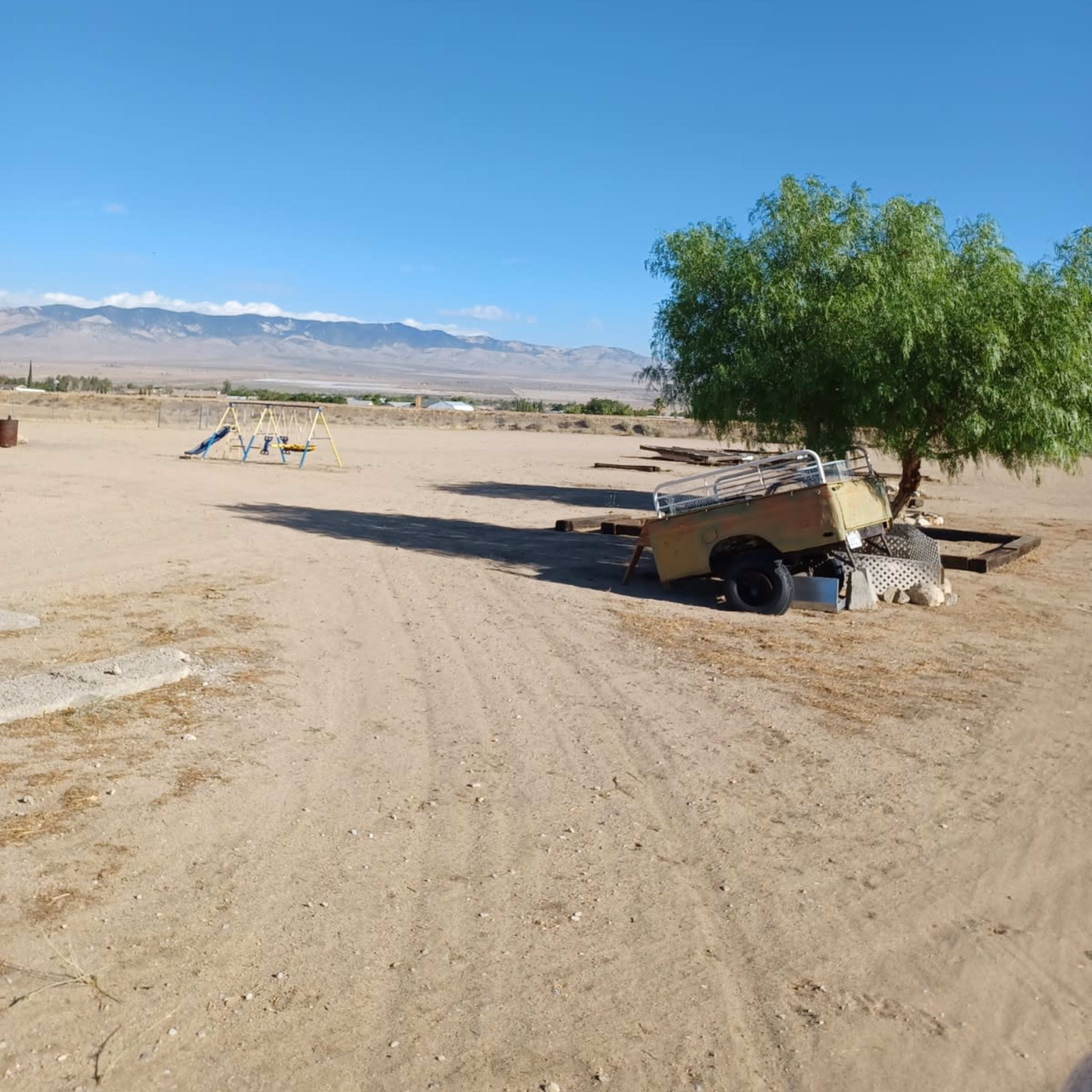 A barren landscape with a parked trailer near a small tree, swings in the background, and mountains in the distance under a clear blue sky.
