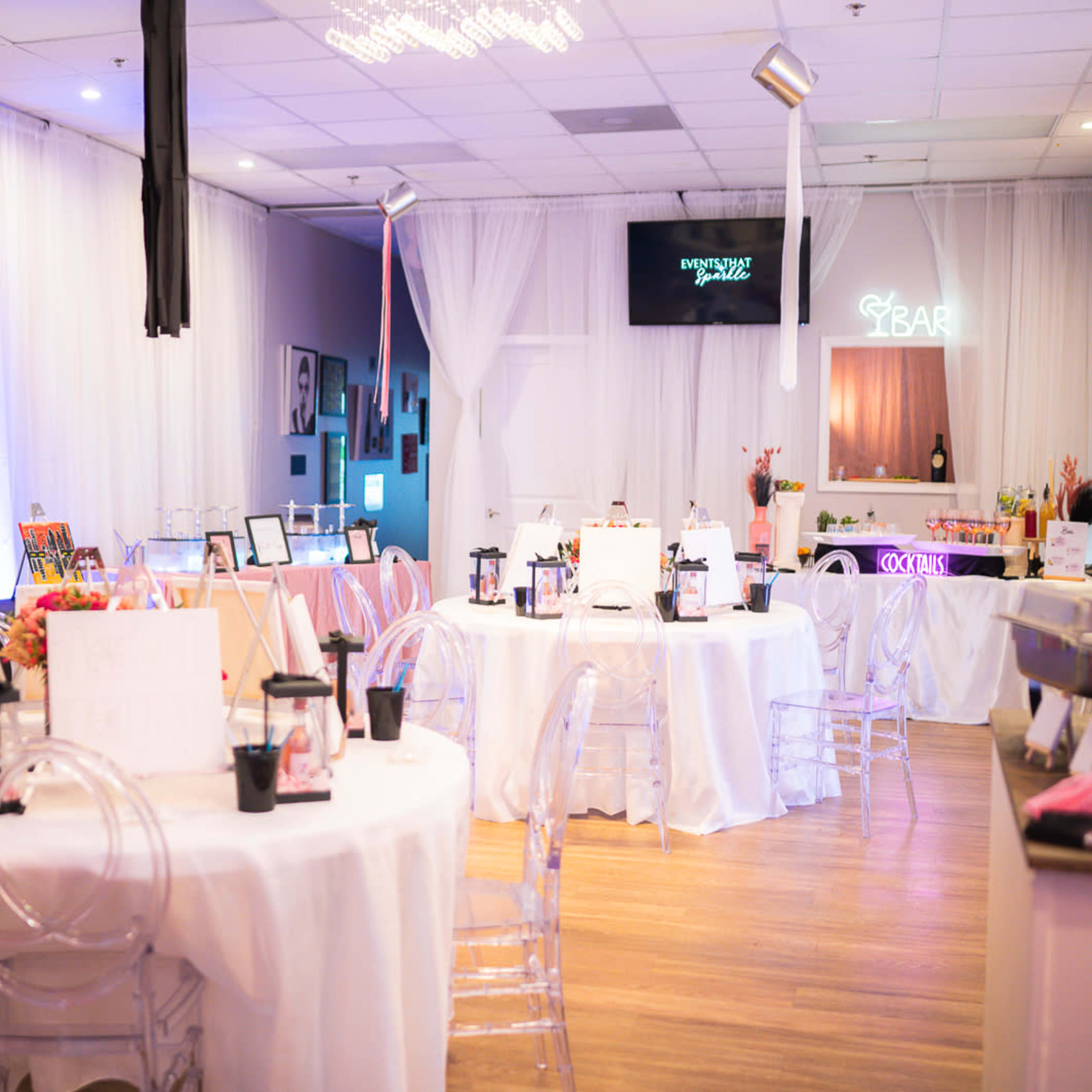 A decorated event space with several round tables set for a gathering, featuring white tablecloths, chairs, and a bar area in the background.