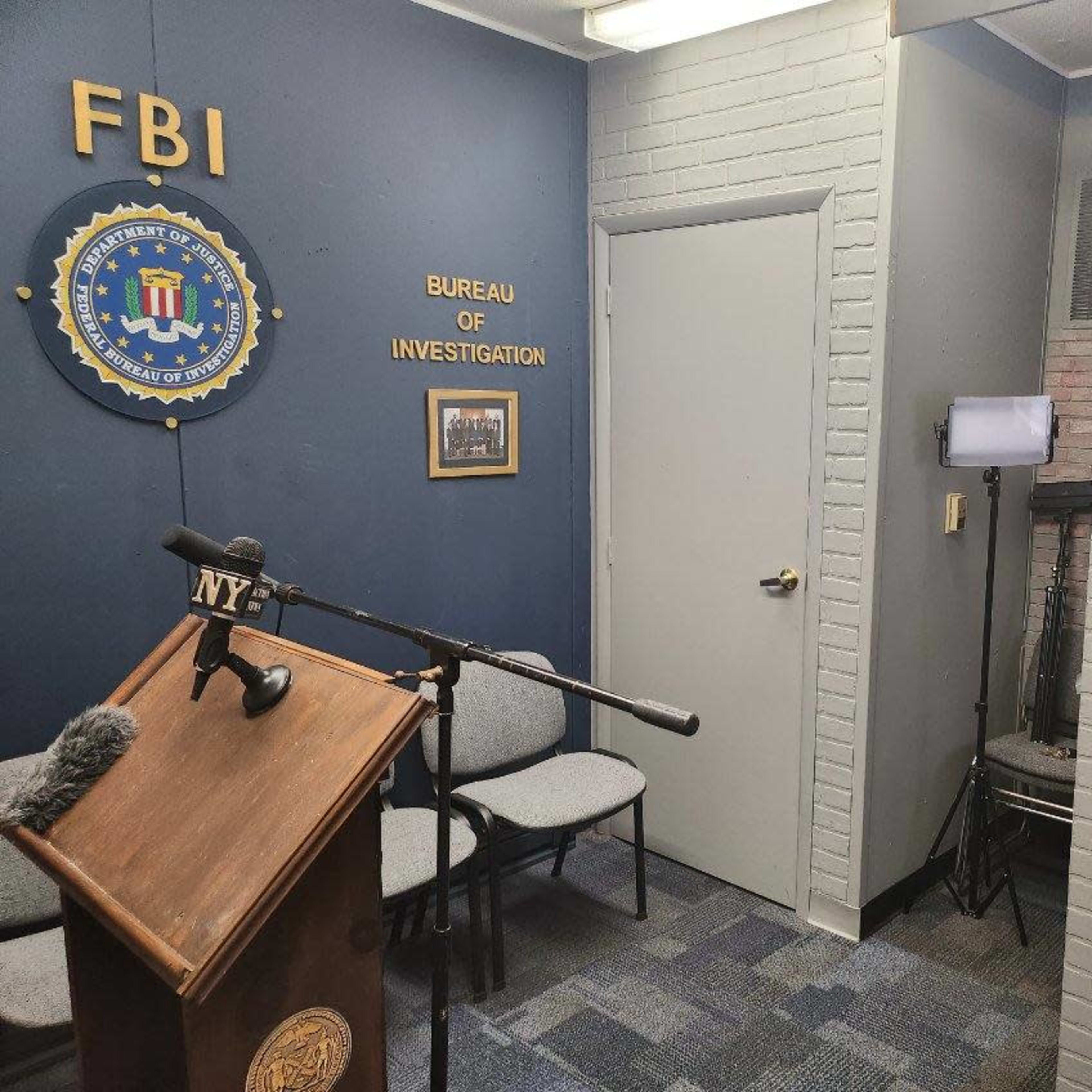 A small room with a podium featuring a microphone, chairs arranged in front of it, and a blue wall displaying an FBI seal.