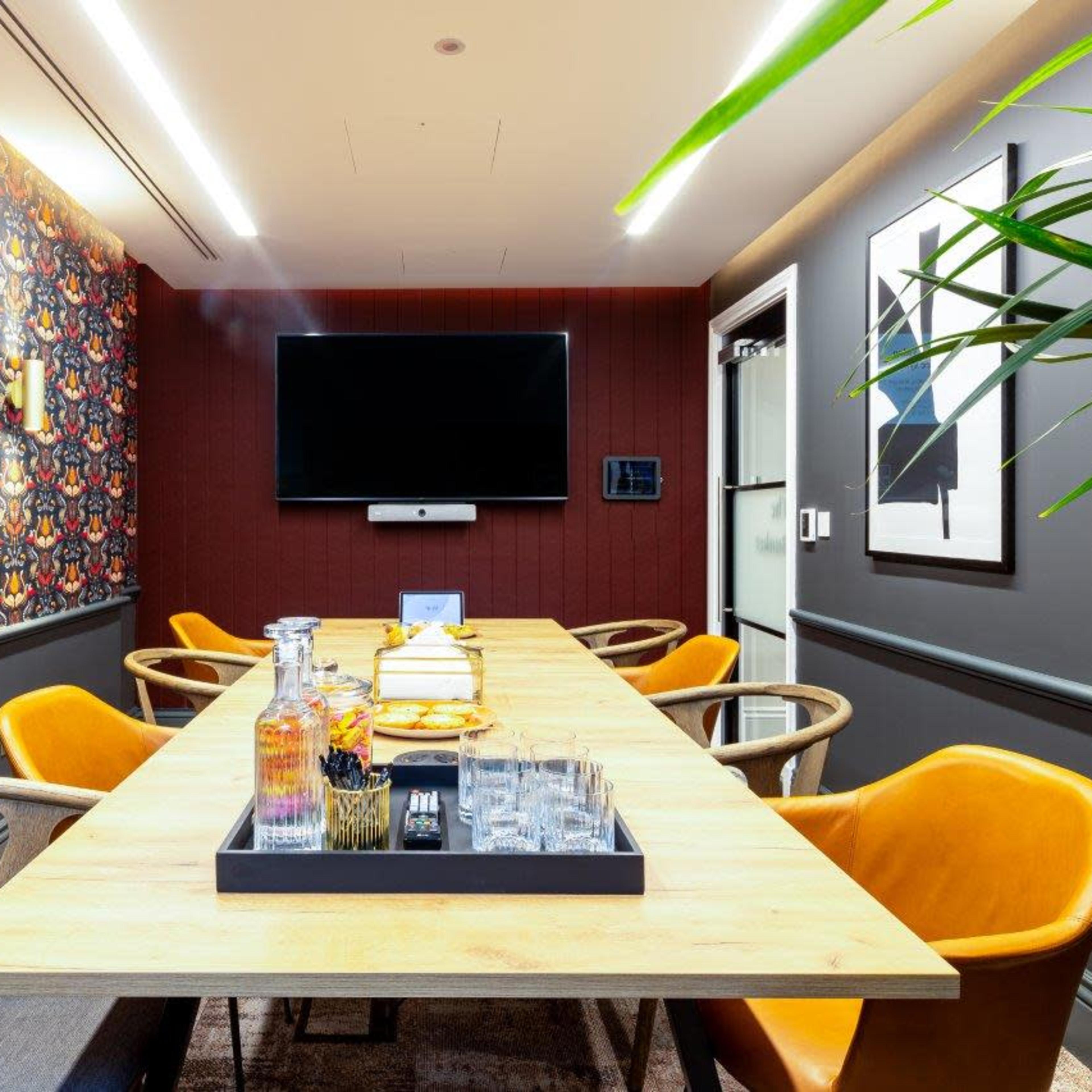 A modern meeting room features a long wooden table surrounded by yellow chairs, with a television mounted on the wall and decorative wallpaper.