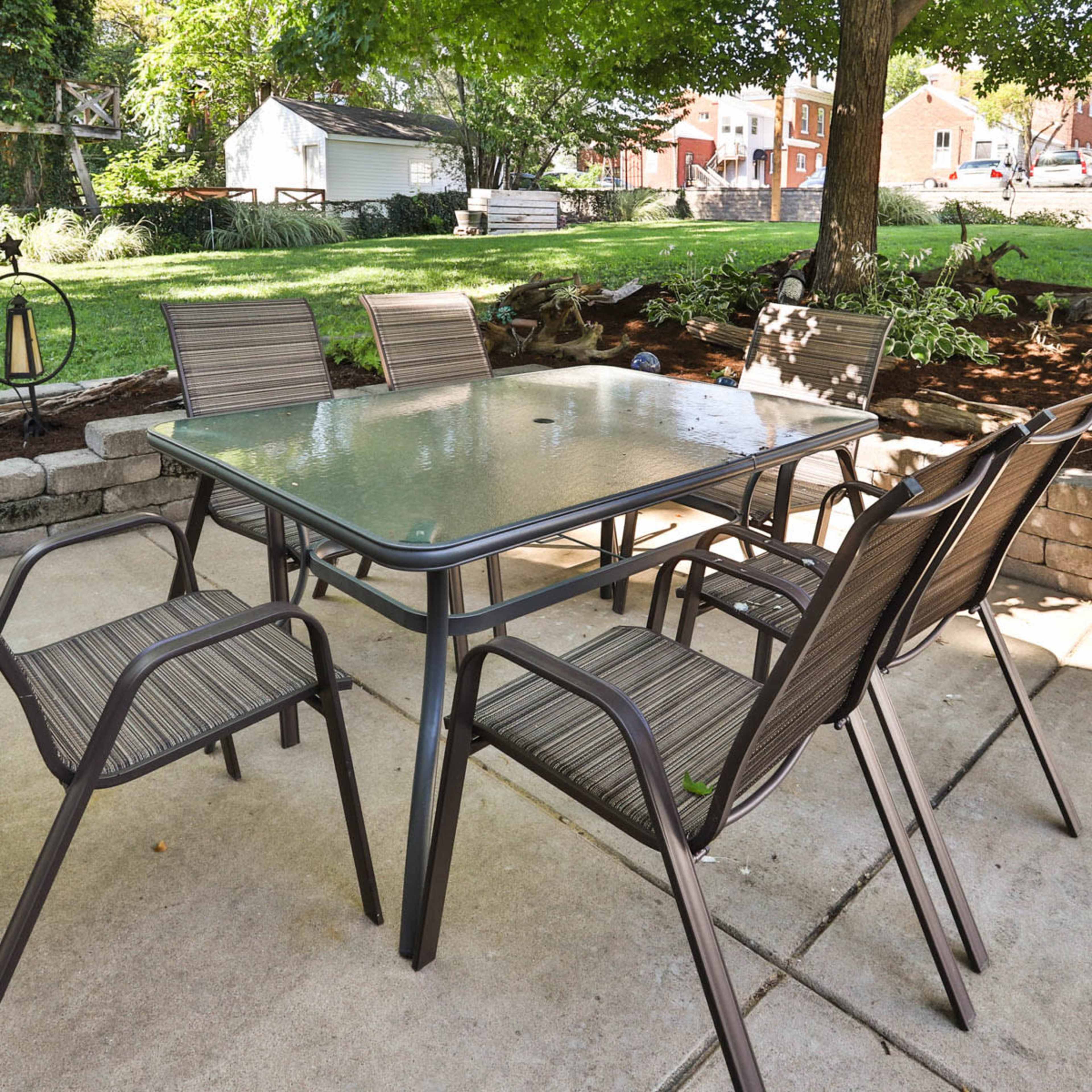 A patio set with a glass table and six chairs is arranged on a concrete surface under a tree in a backyard.