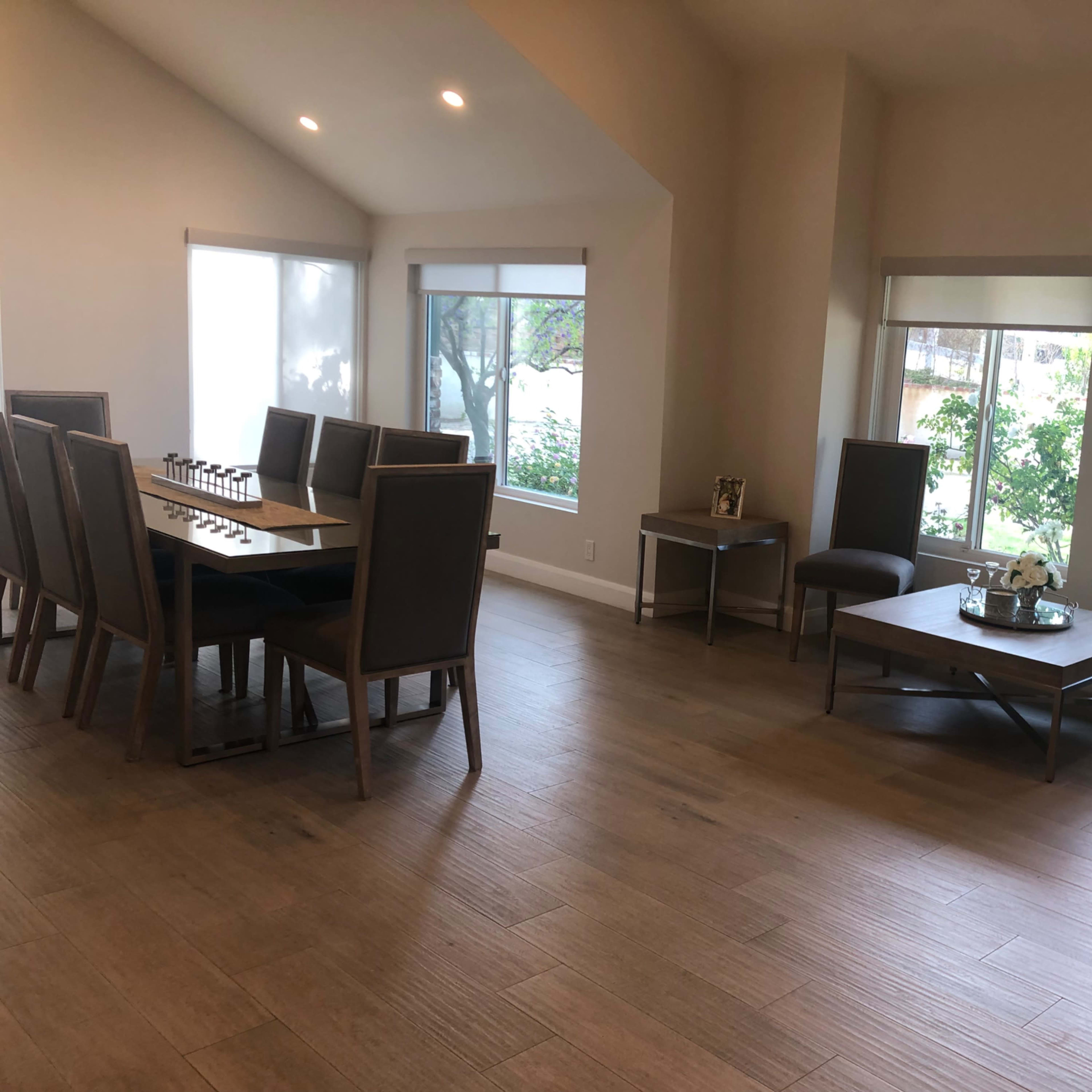 A spacious room features a long table with chairs on one side and several smaller tables with chairs on the other side, all illuminated by natural light from large windows.