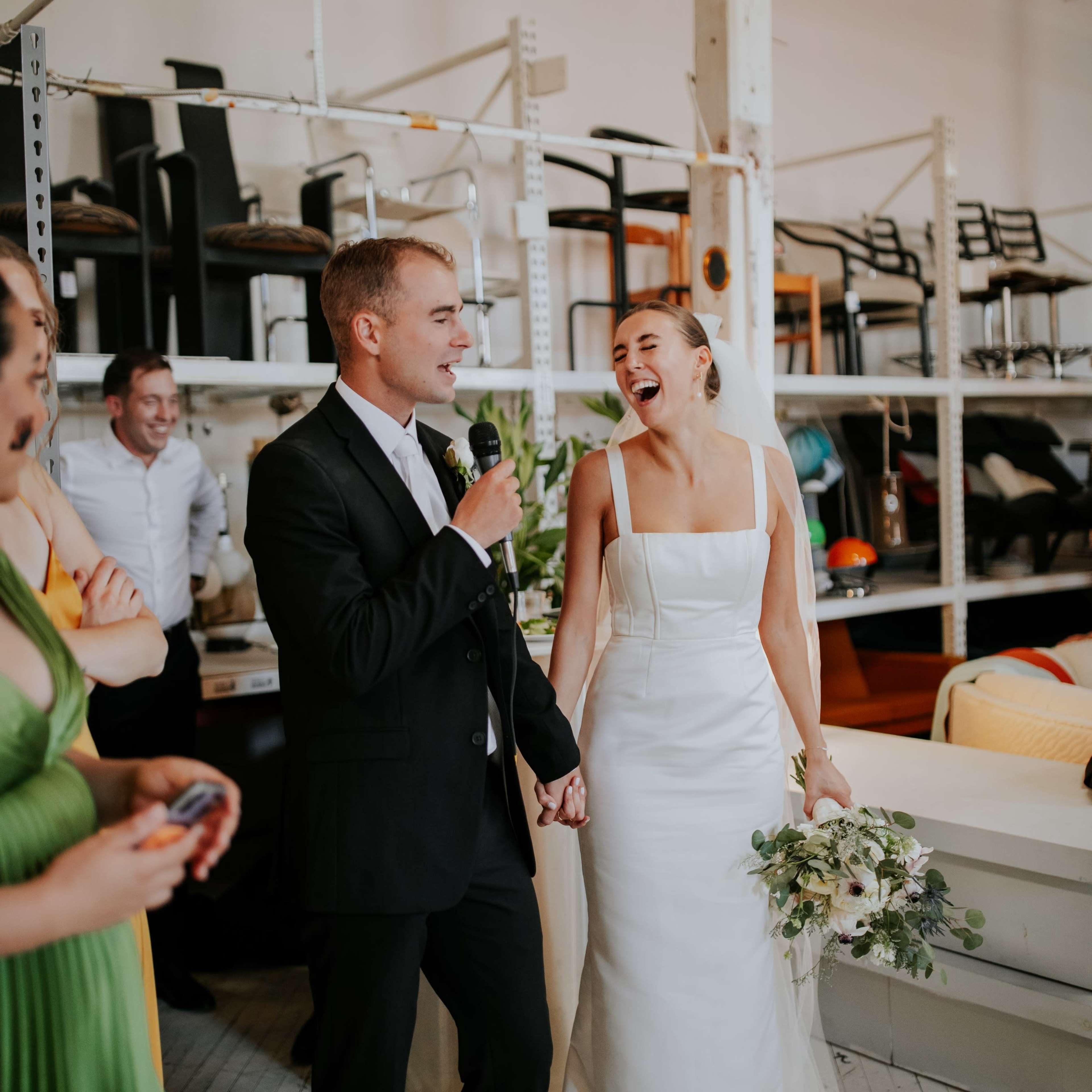 A man in a suit speaks into a microphone while holding hands with a smiling woman in a white wedding dress, surrounded by guests in a bright, industrial-style venue.